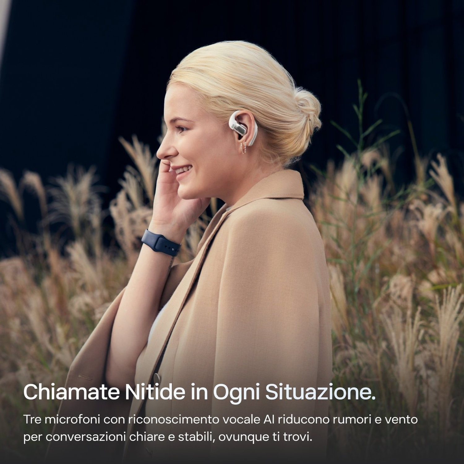 Una donna bionda indossa auricolari e un cappotto beige, sorridendo all'aperto.