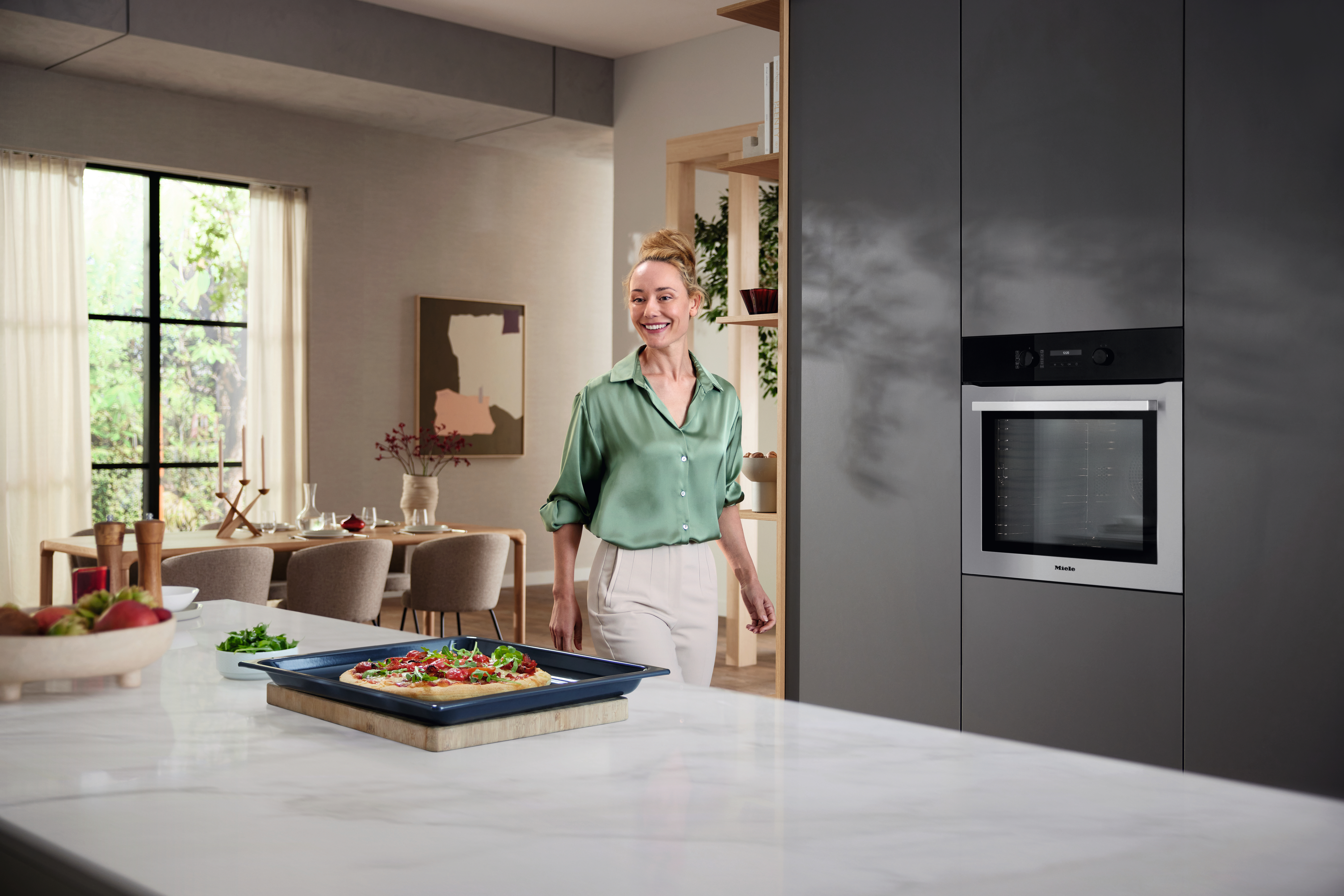Eine Frau in einem grünen Hemd steht in einer modernen Küche mit einem Backofen und einem Esstisch.