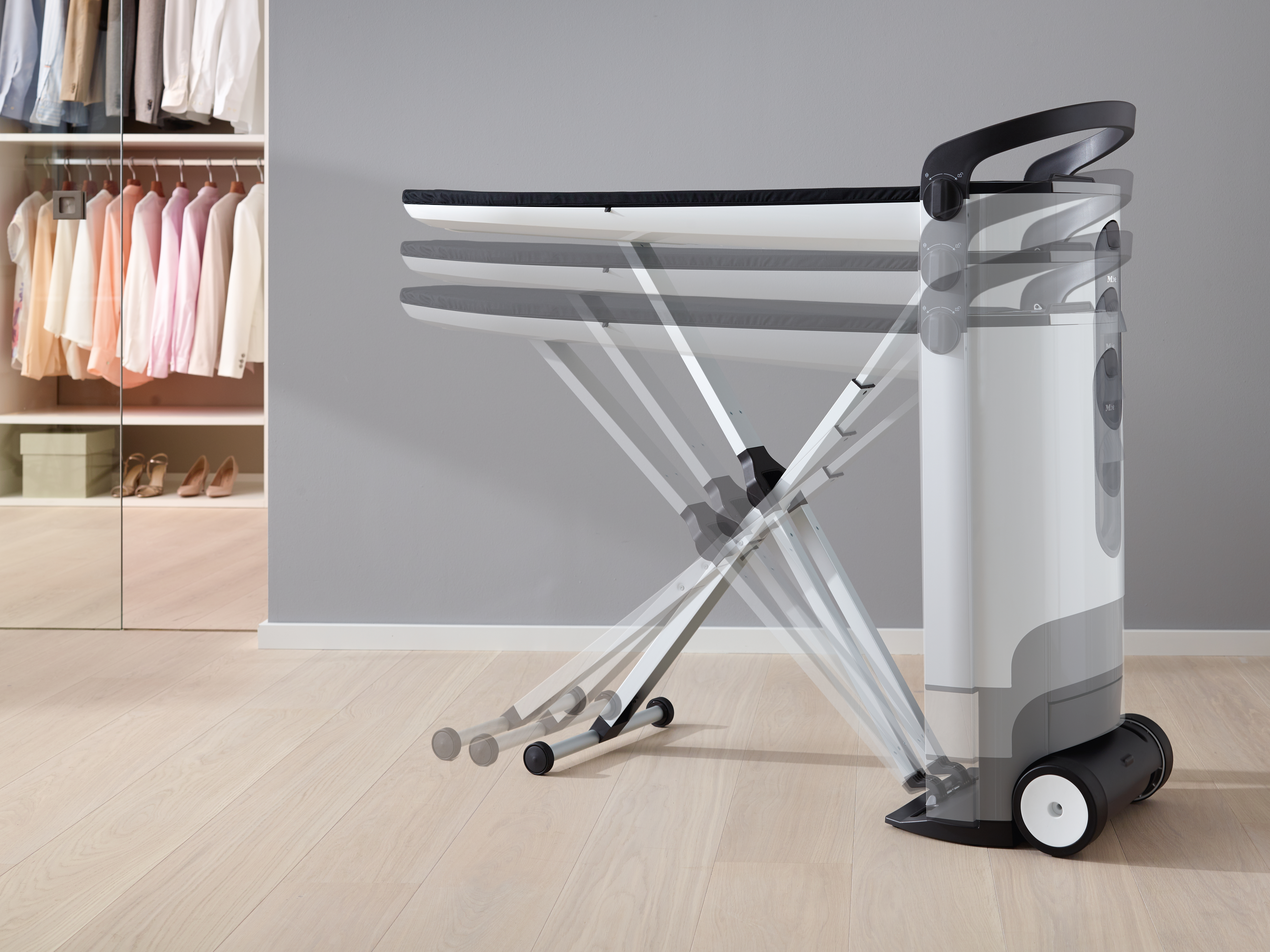 Ein Bügelbrett mit Rollen und einem Kleiderschrank voller Kleidung im Hintergrund.