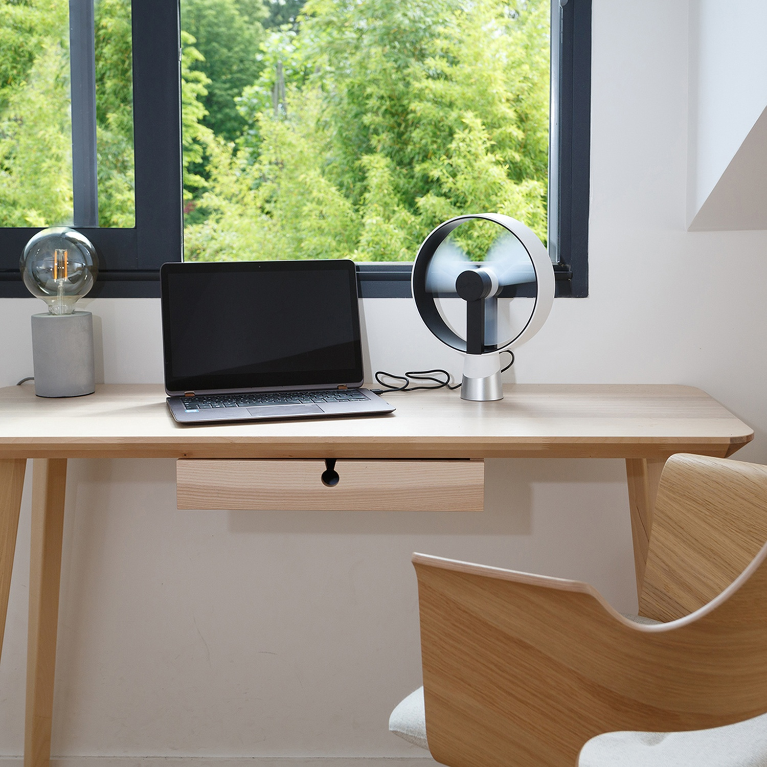 Bureau met laptop, lamp en ventilator bij een raam met uitzicht op groene bomen.