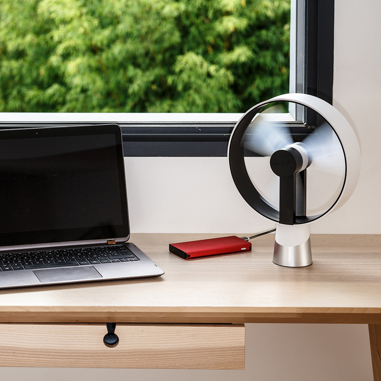 Een laptop, rode powerbank en ventilator op een houten bureau bij een raam.