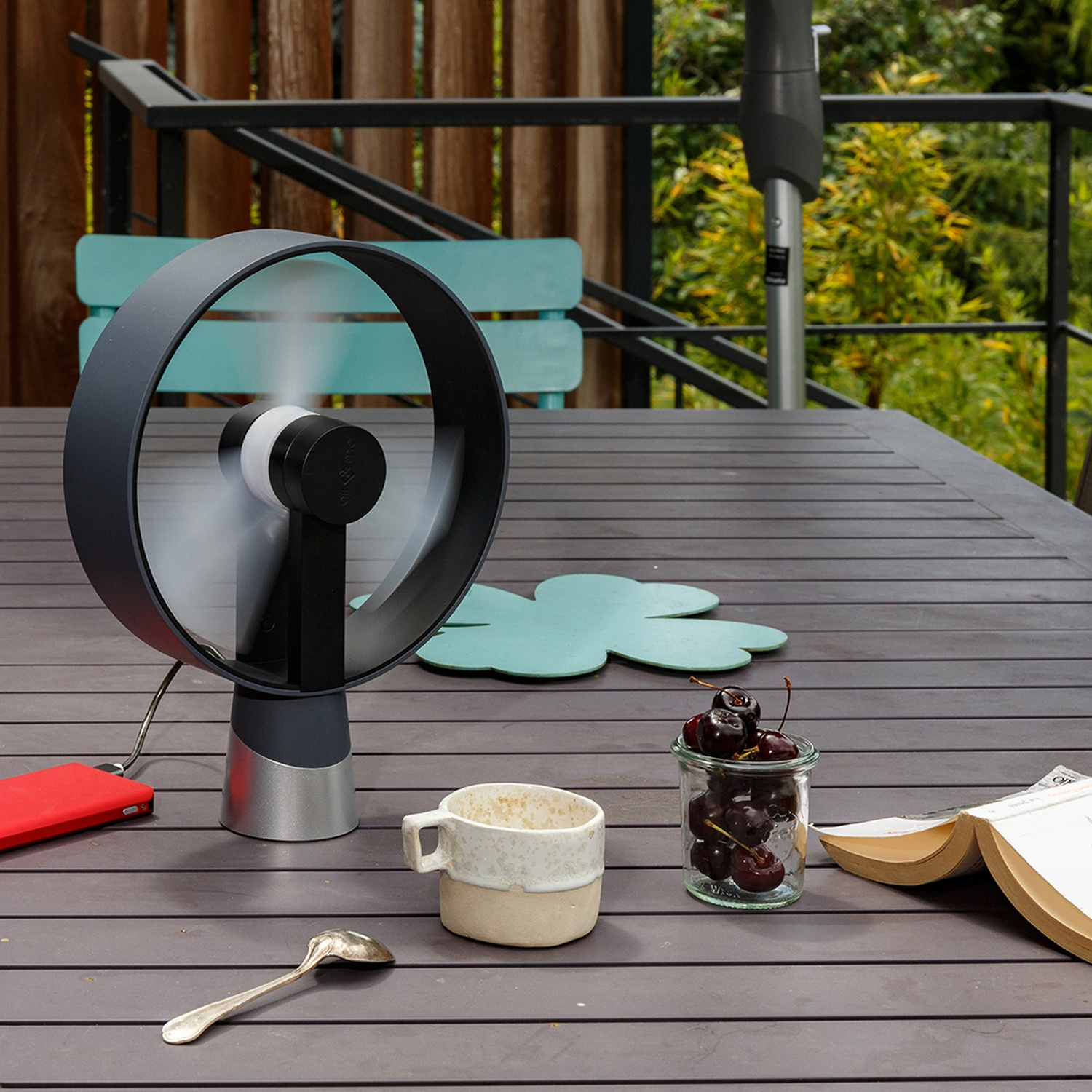 Draagbare ventilator, rode telefoon, kopje, kersen in pot en open boek op houten tafel.