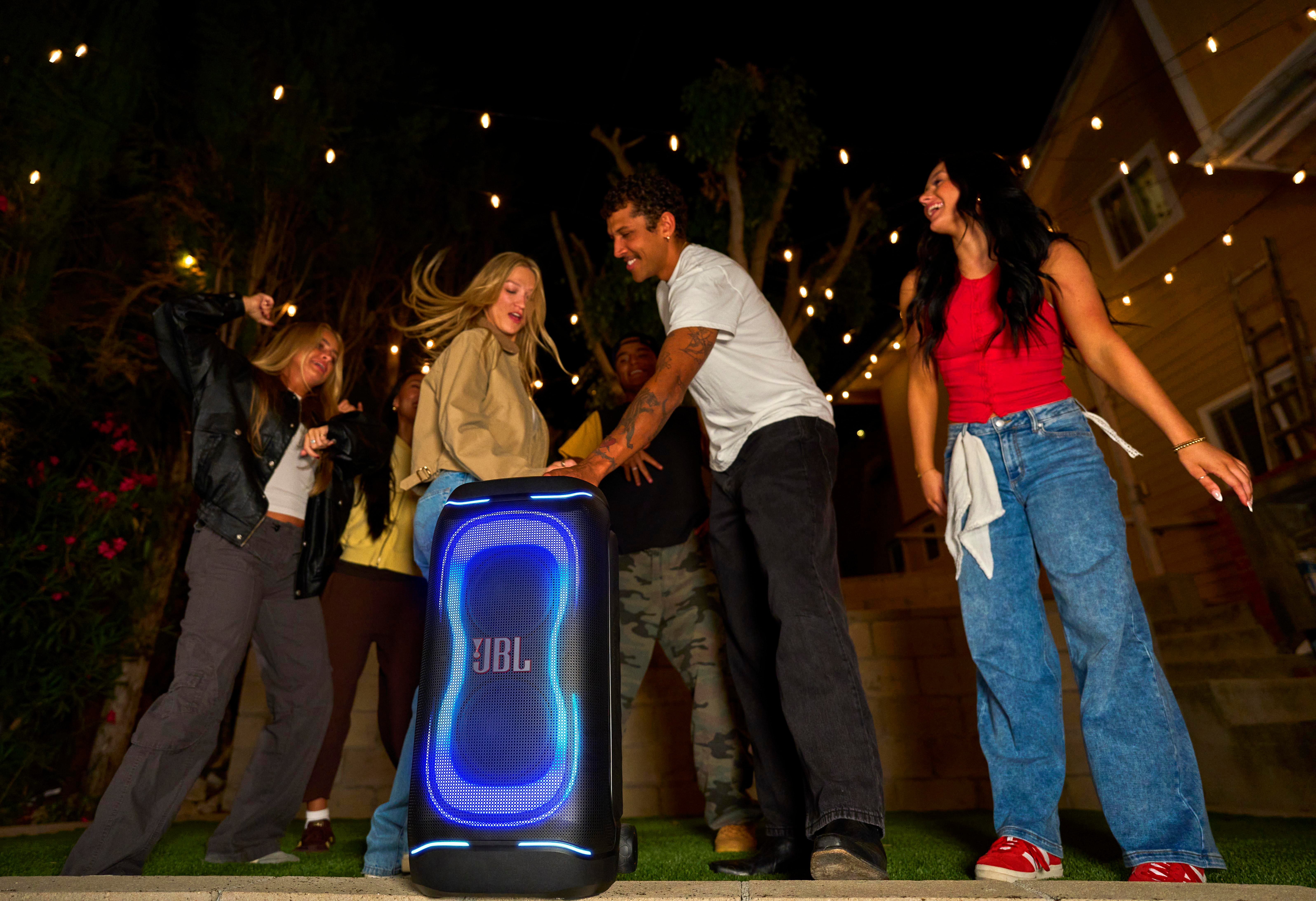 Freunde tanzen um einen leuchtenden blauen JBL-Lautsprecher bei einer nächtlichen Gartenparty.