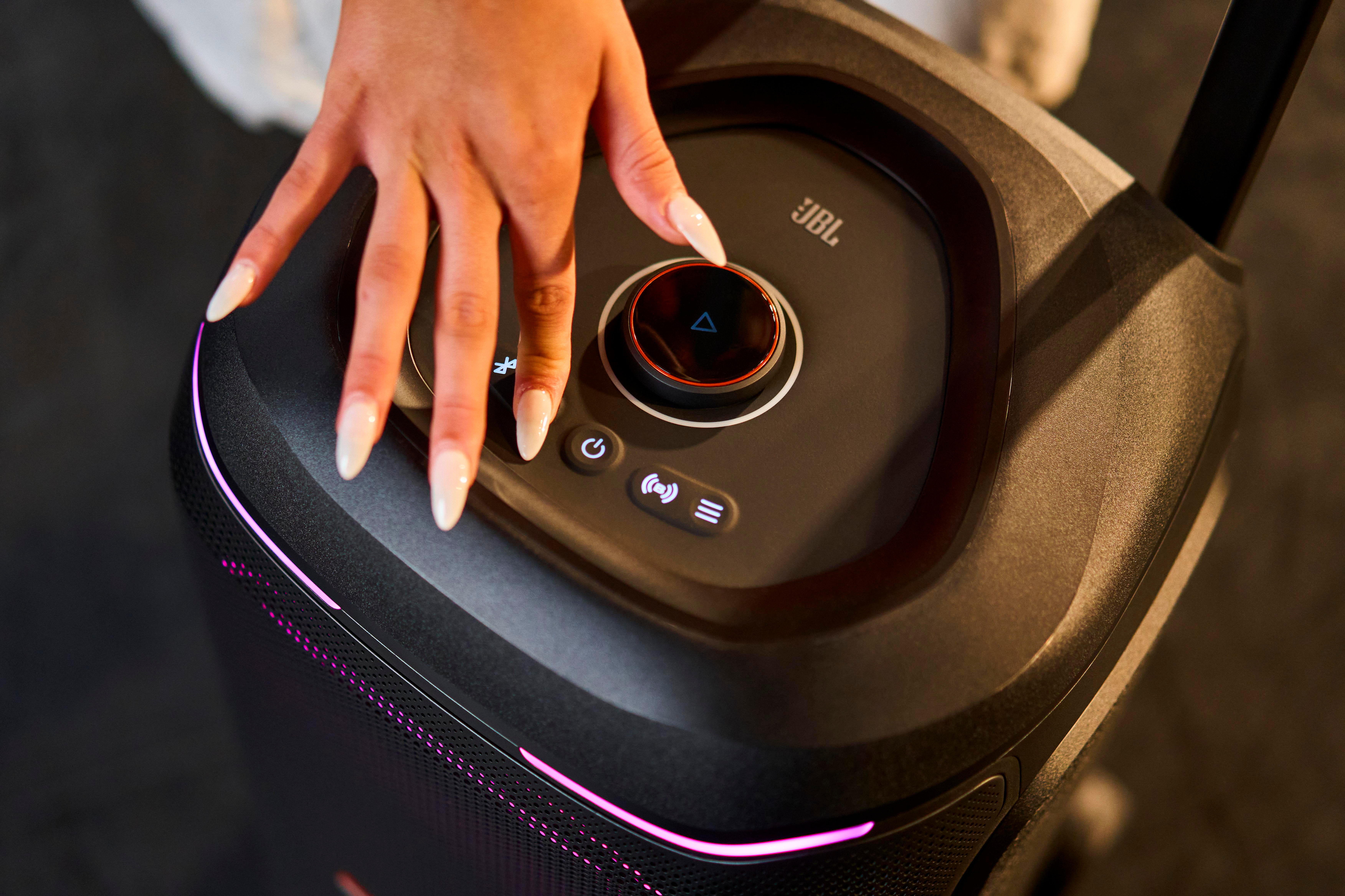 Eine Hand mit langen Nägeln berührt das Bedienfeld eines schwarzen JBL-Lautsprechers mit pinker Beleuchtung.