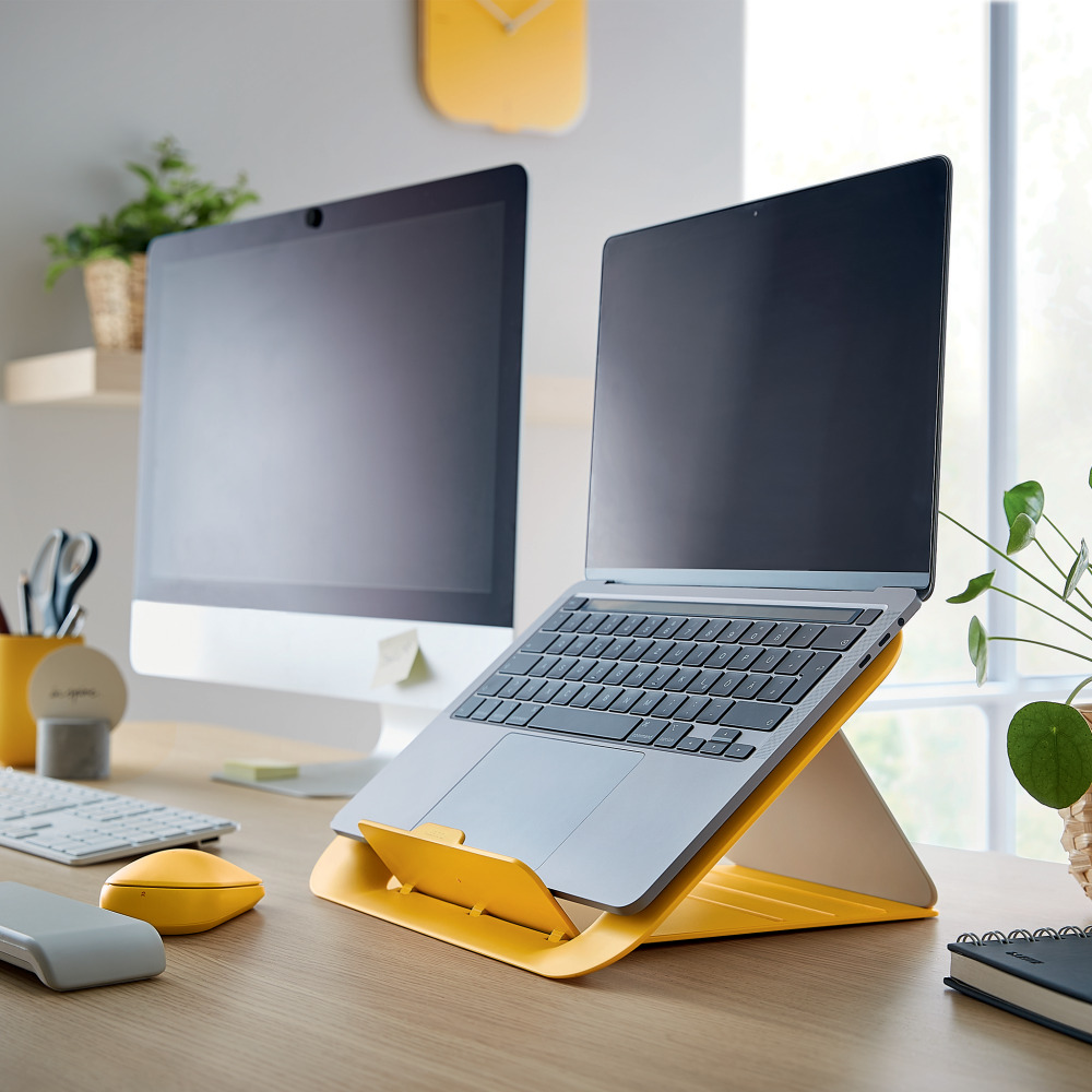 Laptop sárga állványon, mellette számítógép monitor az asztalon.