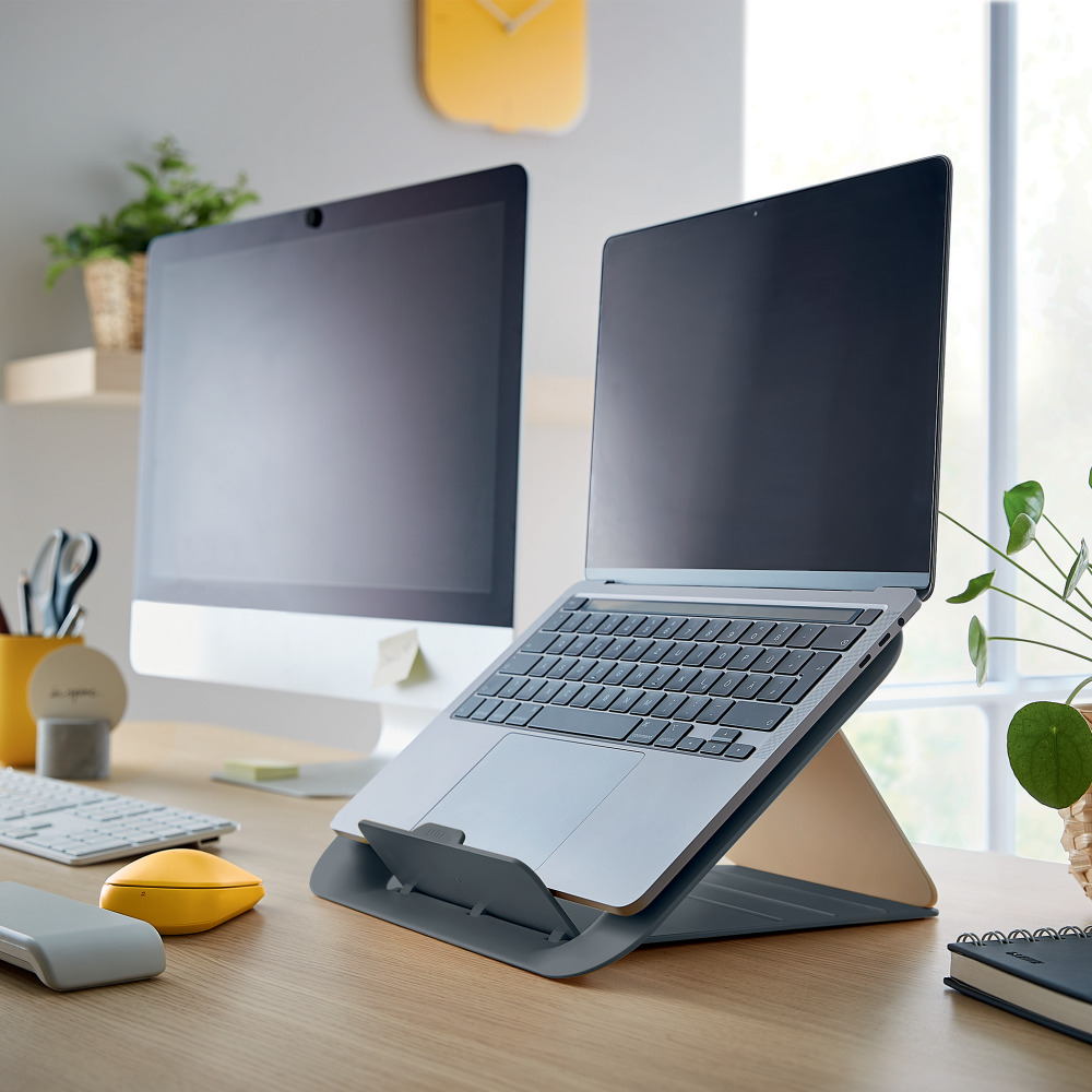 Laptop állványon, számítógép monitorral és sárga egérrel az asztalon.
