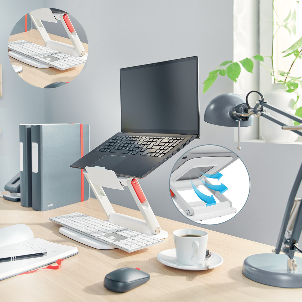 Egy laptop állványon van egy billentyűzet fölött. Közelben egér, kávé és jegyzetfüzet.