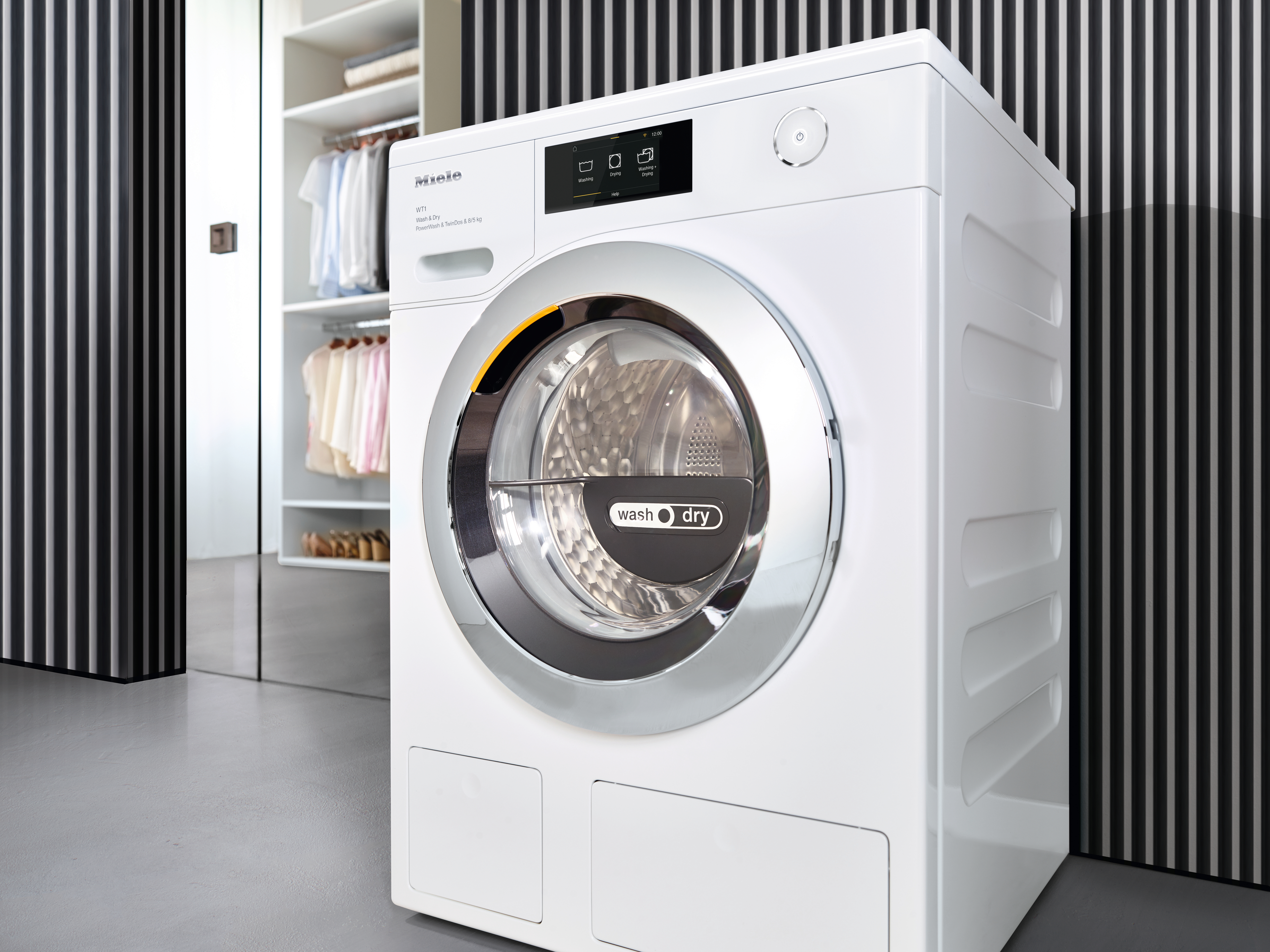 Weiße Miele Waschmaschine mit silbernem Rand und Display. Daneben ein offener Schrank mit Kleidung.
