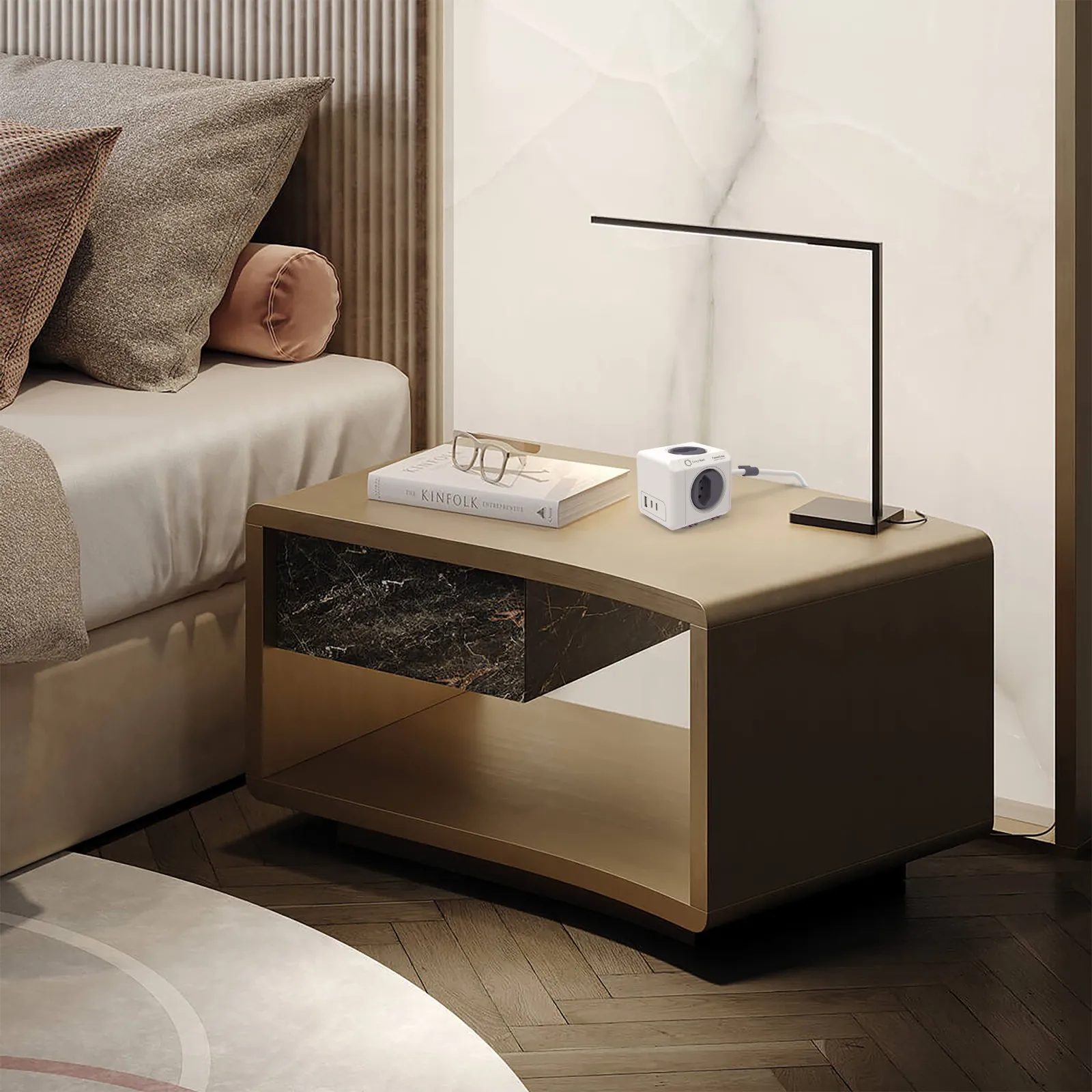 Ein Nachttisch mit Buch, Lampe und Steckdosenwürfel neben einem Bett.