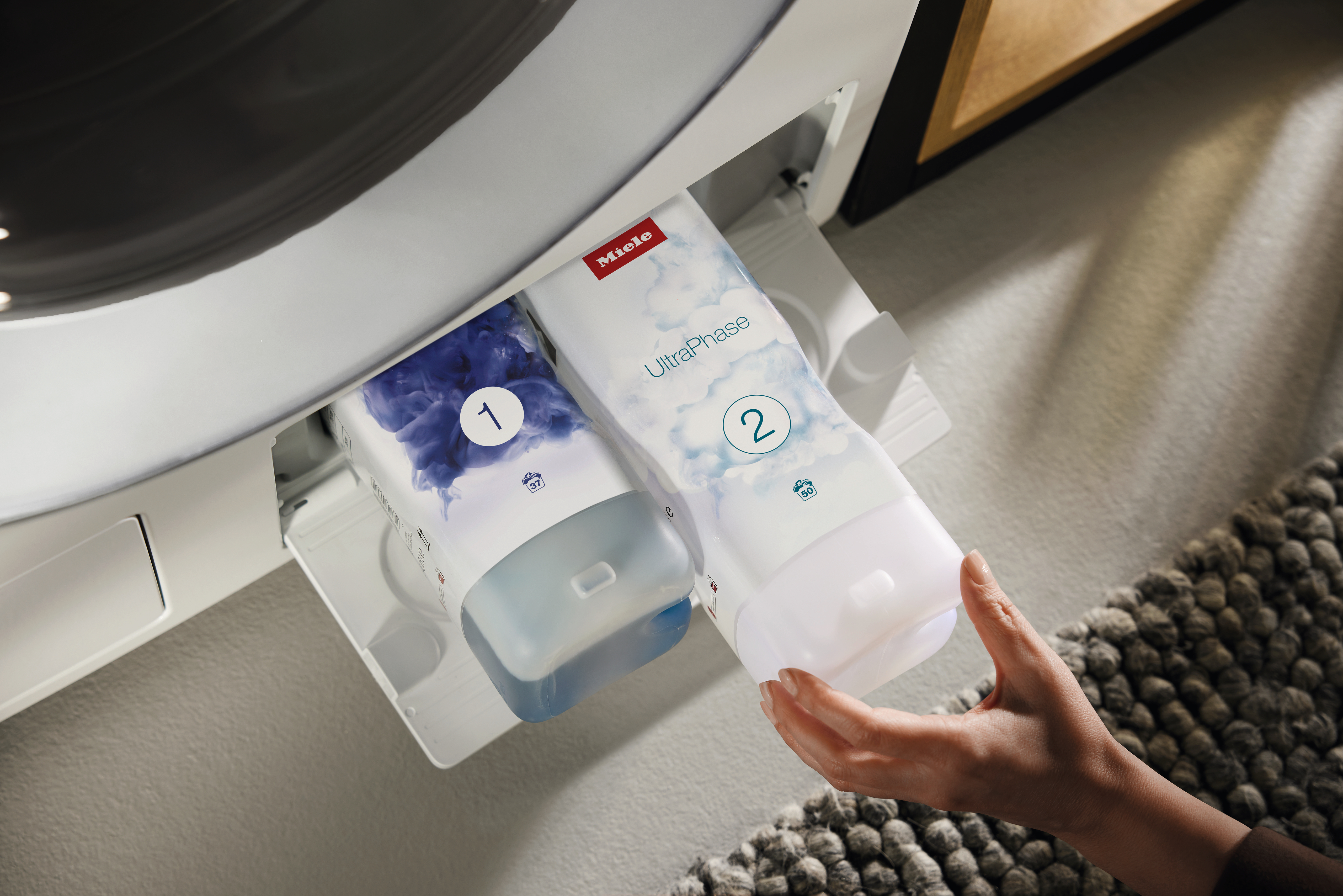 Eine Hand schiebt zwei Miele UltraPhase Waschmittelkapseln in eine Waschmaschine.
