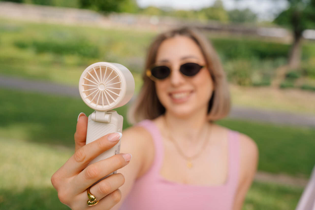 Egy nő mosolyog, miközben egy kis ventilátort tart. Napszemüveget visel.