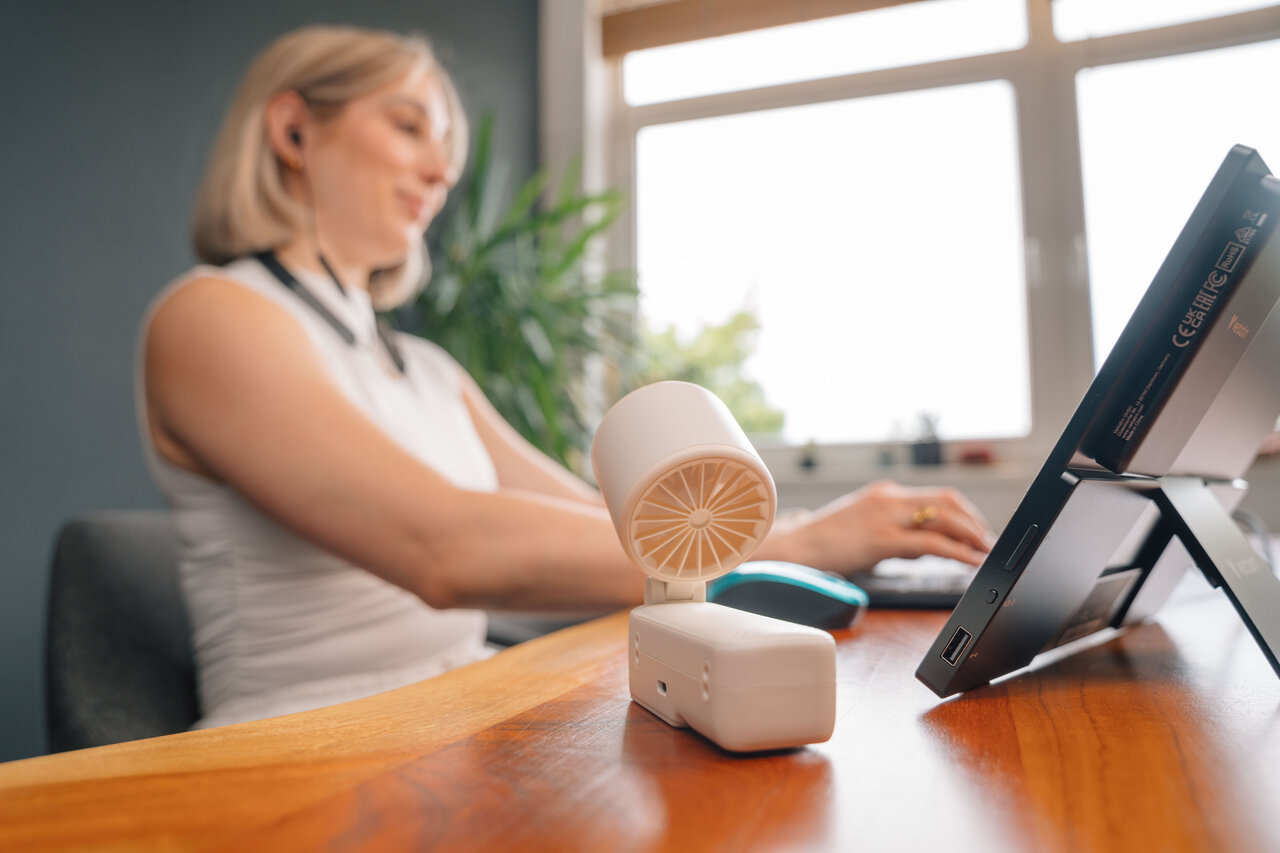 Egy nő laptopon dolgozik, egy kis ventilátor az asztalon.