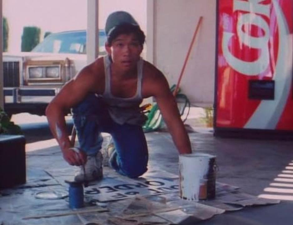 Junger Mann im Tanktop und Jeans bemalt einen blauen Eimer. Coca-Cola-Automat in der Nähe.