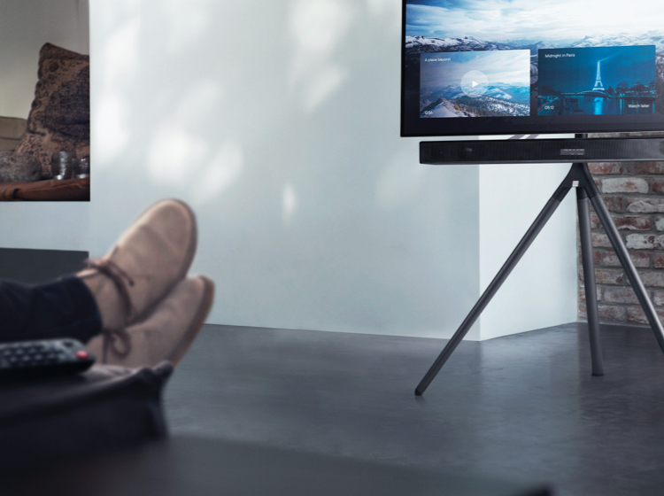 Persona relajándose, pies sobre mesa con mando, viendo TV en soporte trípode.