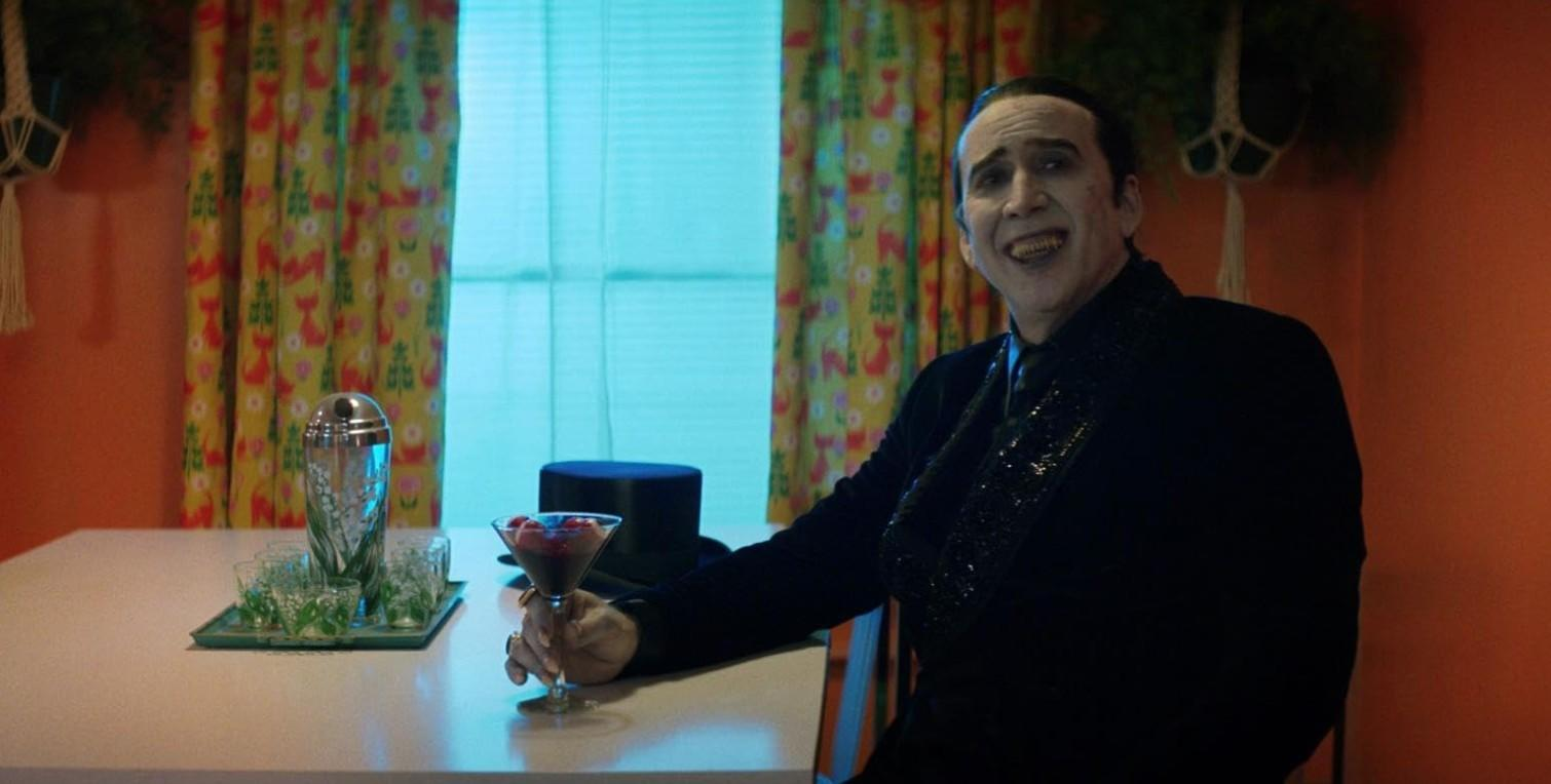 Mann mit weißem Make-up hält ein Glas Cocktail in einem Raum mit orangefarbenen Wänden.