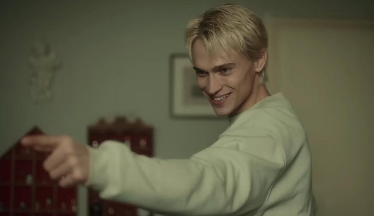 Ein Mann mit blonden Haaren lächelt und zeigt nach links. Er trägt ein hellgrünes Sweatshirt.