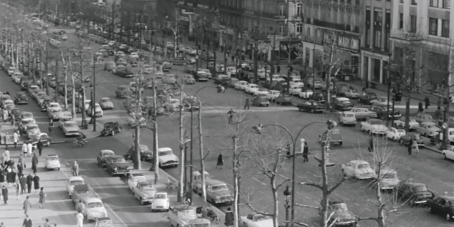 Eine belebte Straßenszene mit vielen Autos und Fußgängern, Bäumen und Gebäuden.