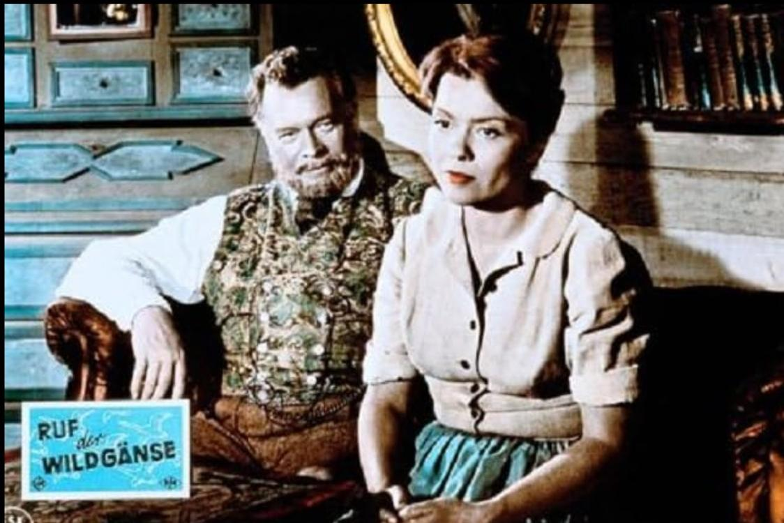 Mann mit Bart und Frau sitzen nebeneinander. Filmplakat im Vordergrund.