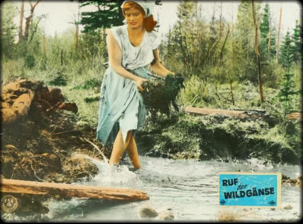 Frau mit Kopftuch in einem blauen Kleid watet durch Wasser und hält Schlamm.