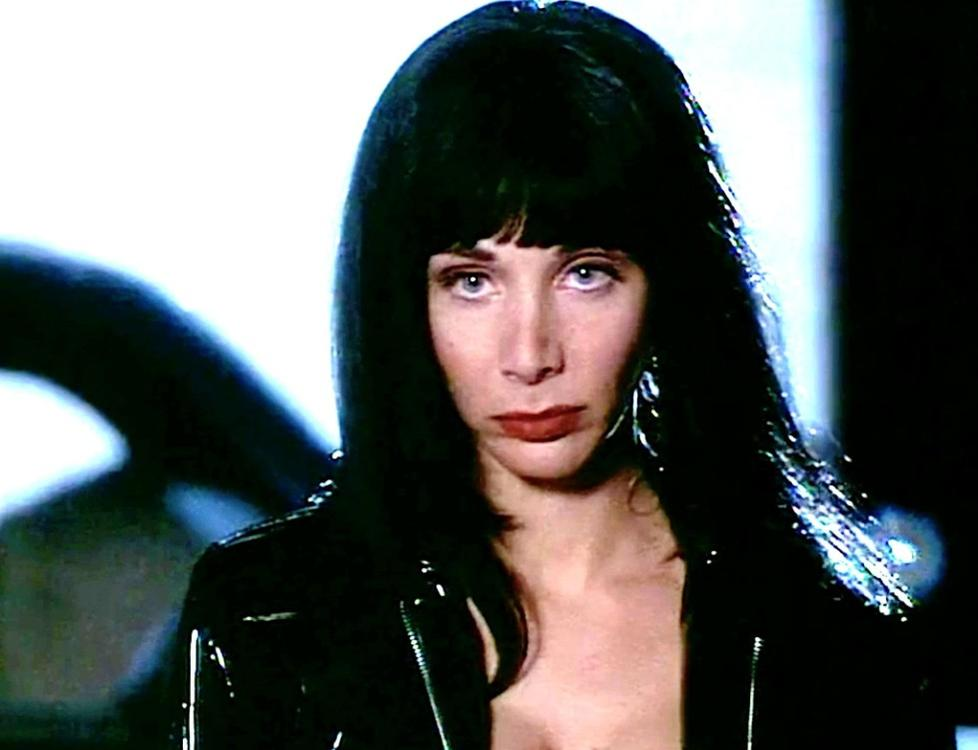 Eine Frau mit dunklem Haar und Make-up trägt eine schwarze Jacke. Ihr Ausdruck ist ernst.