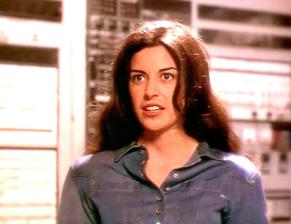 Eine Frau mit braunen Haaren trägt ein blaues Jeanshemd und blickt überrascht.