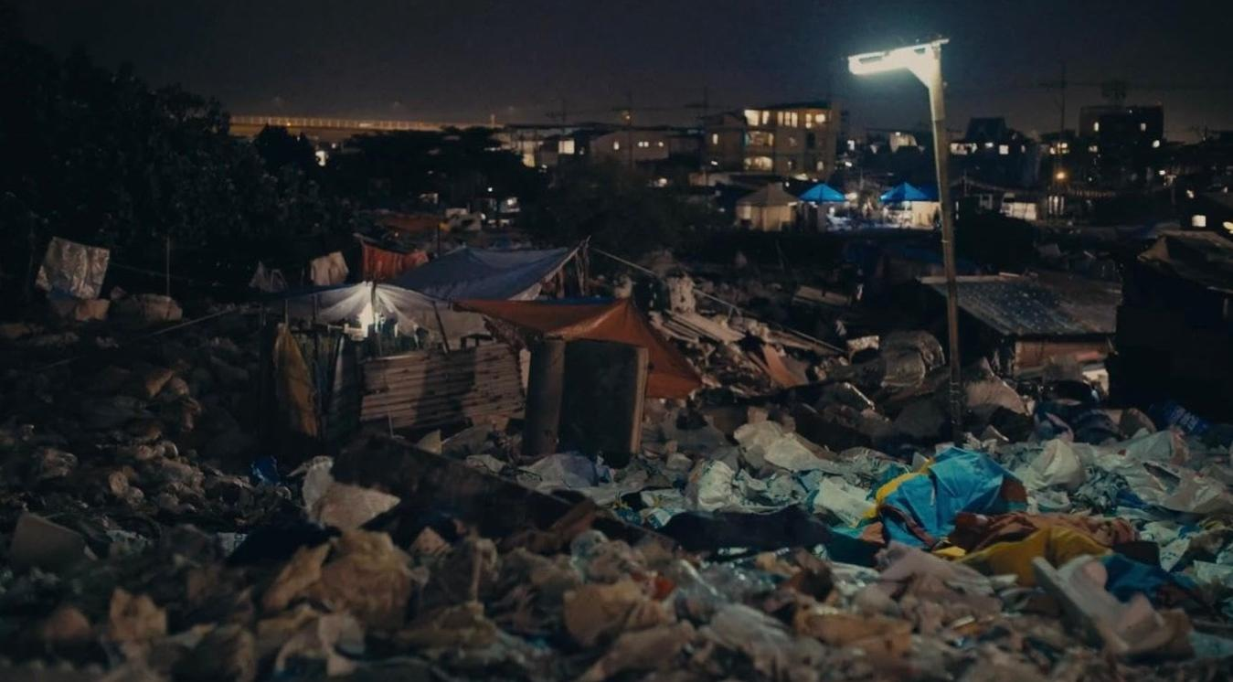 Nächtliche Szene eines Slums mit Behelfszelten, Müll und einer Straßenlaterne, die den Bereich beleuchtet.