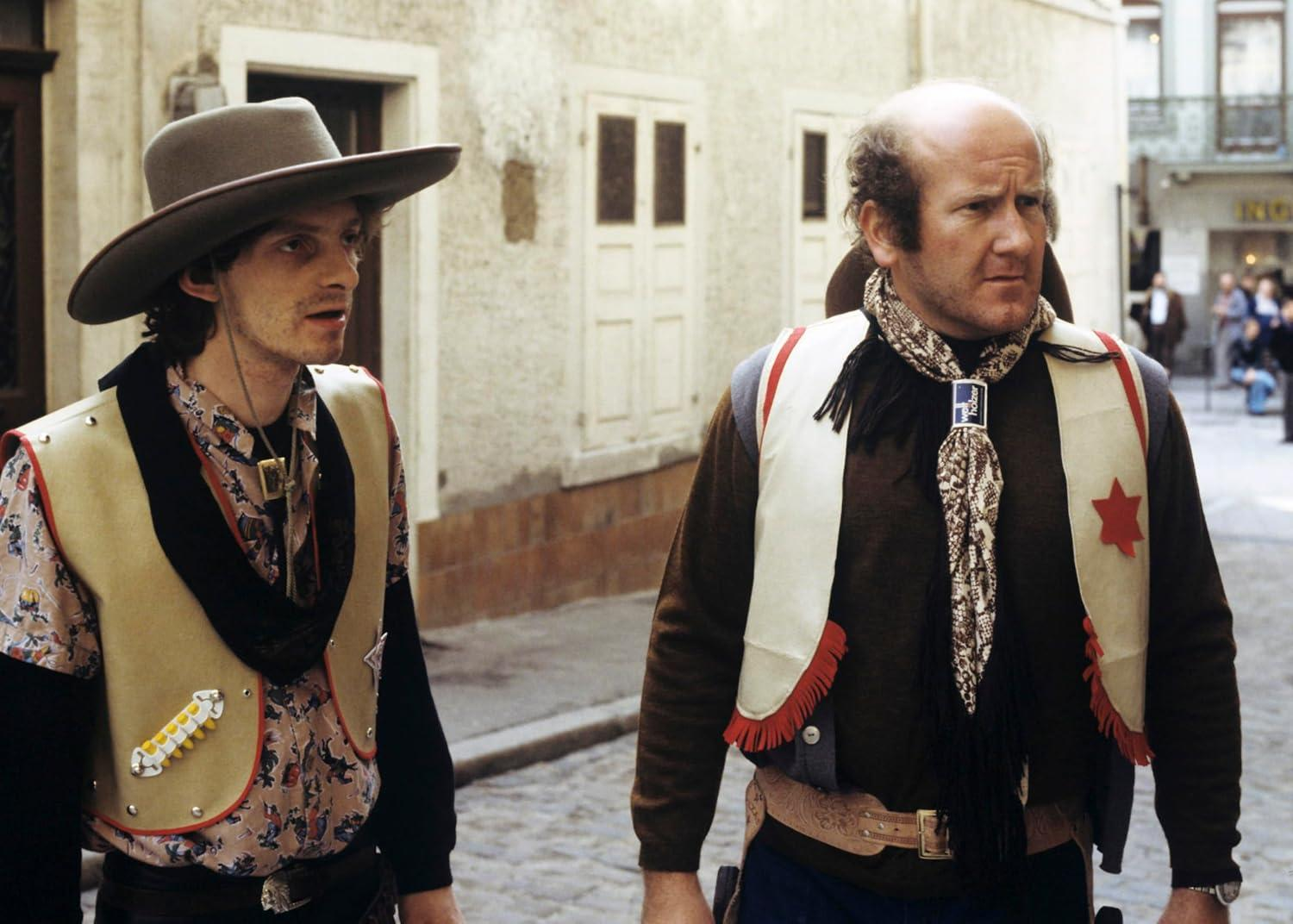 Zwei Männer in Cowboy-Kleidung mit Westen und Hüten gehen auf einer gepflasterten Straße.