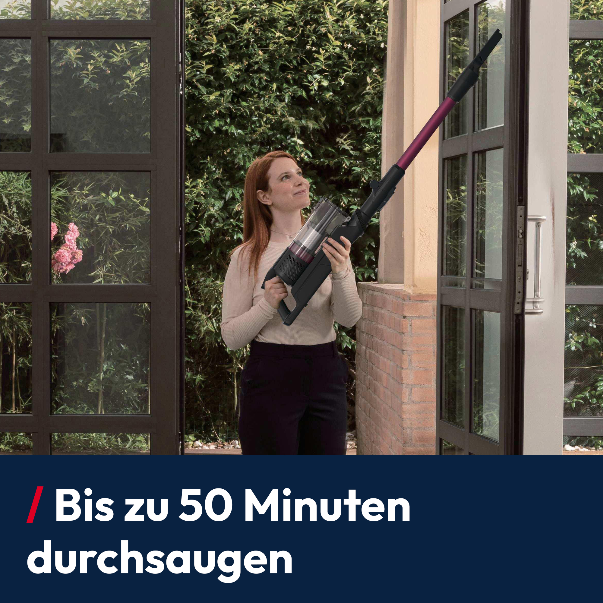 Eine Frau hält einen Stabstaubsauger in einem Innenhof mit viel Grün.