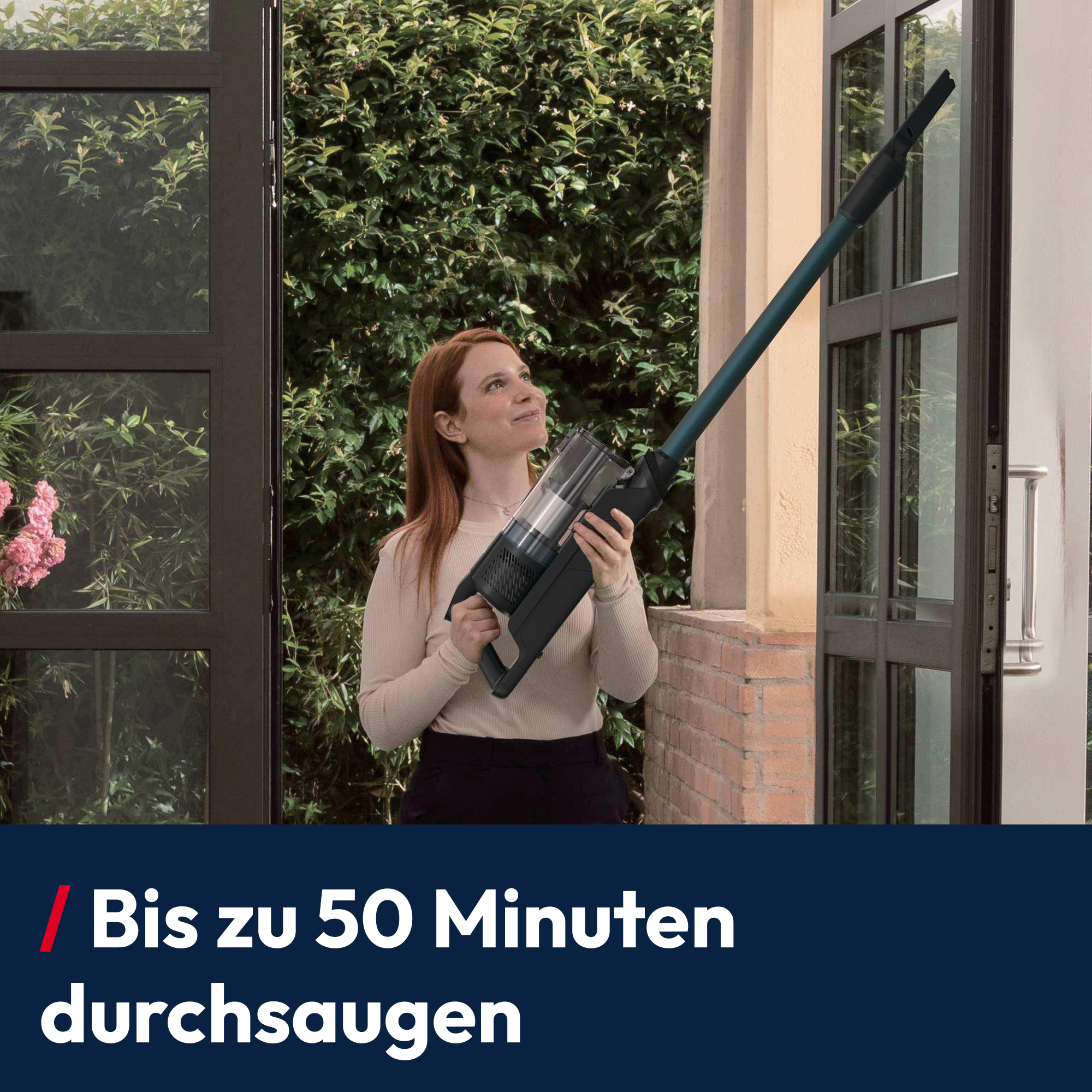 Frau hält Staubsauger vor Glasfront, umgeben von Grünpflanzen.