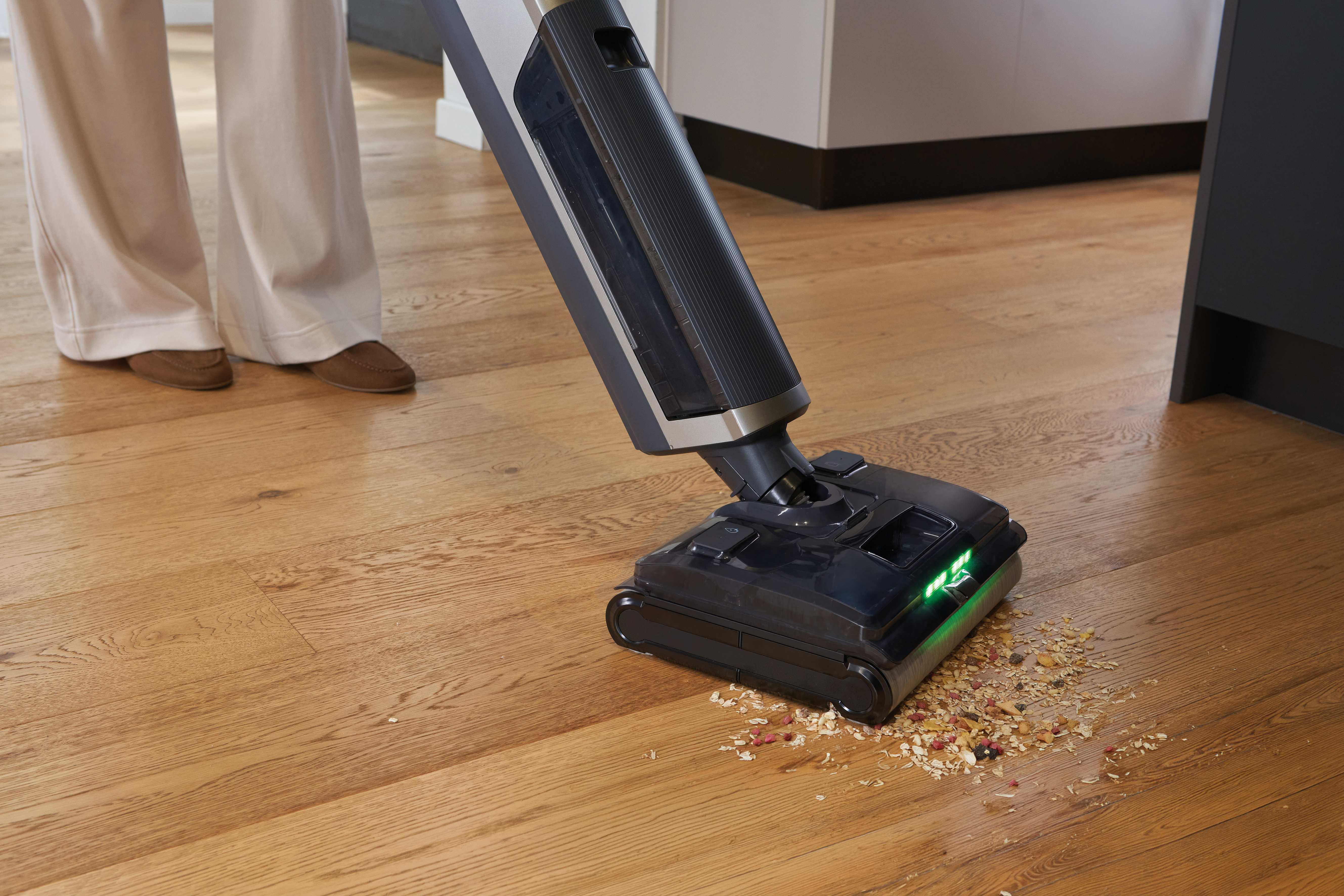 Ein Staubsauger reinigt Holzboden von Essensresten. Grüne Lichter leuchten.