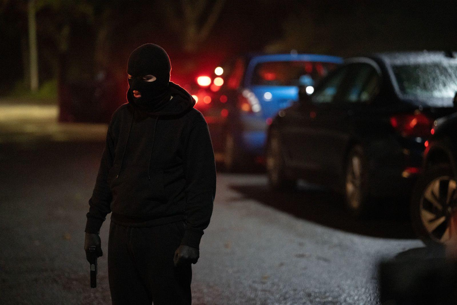Ein Mann mit schwarzer Maske und Kapuze hält eine Pistole in der Hand.