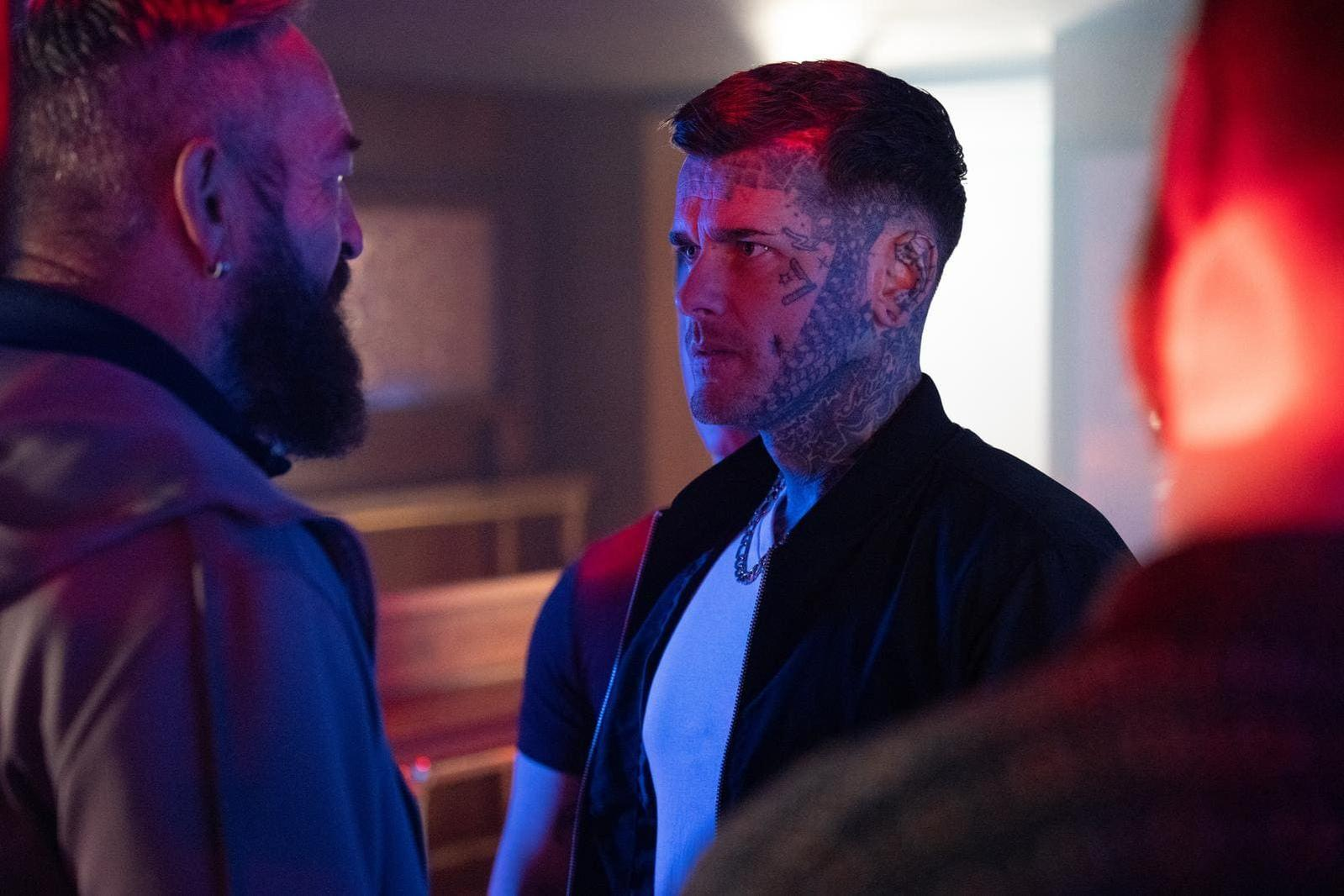 Ein Mann mit Gesichts-Tattoos spricht mit einem anderen Mann in einem Raum mit rotem und blauem Licht.