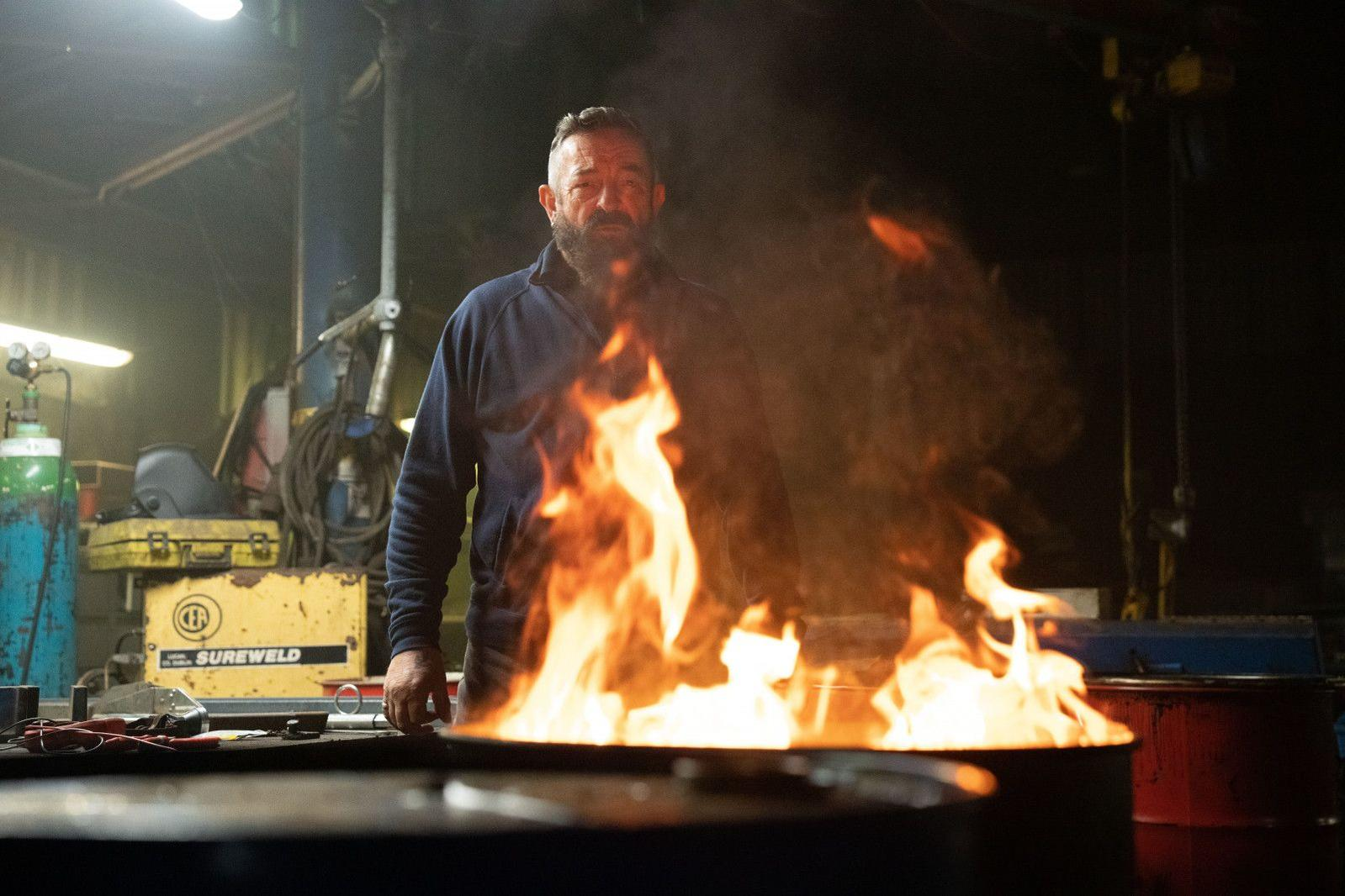 Ein Mann mit Bart steht vor einem brennenden Fass in einer Werkstatt.
