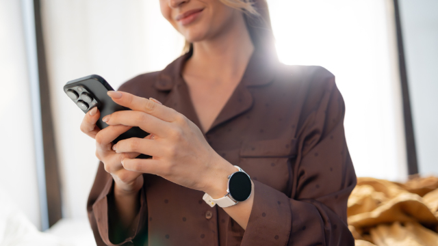 Una donna in pigiama di seta marrone usa uno smartphone e indossa uno smartwatch bianco.