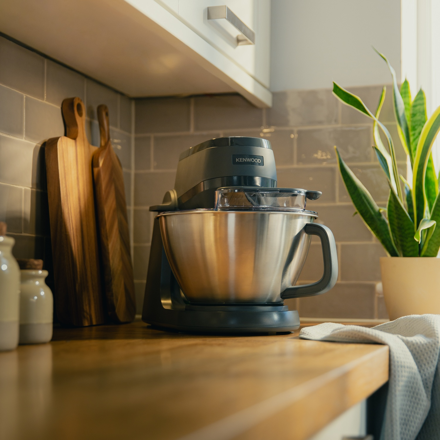 A Kenwood kitchen mixer sits on a countertop next to wooden cutting boards and a plant.