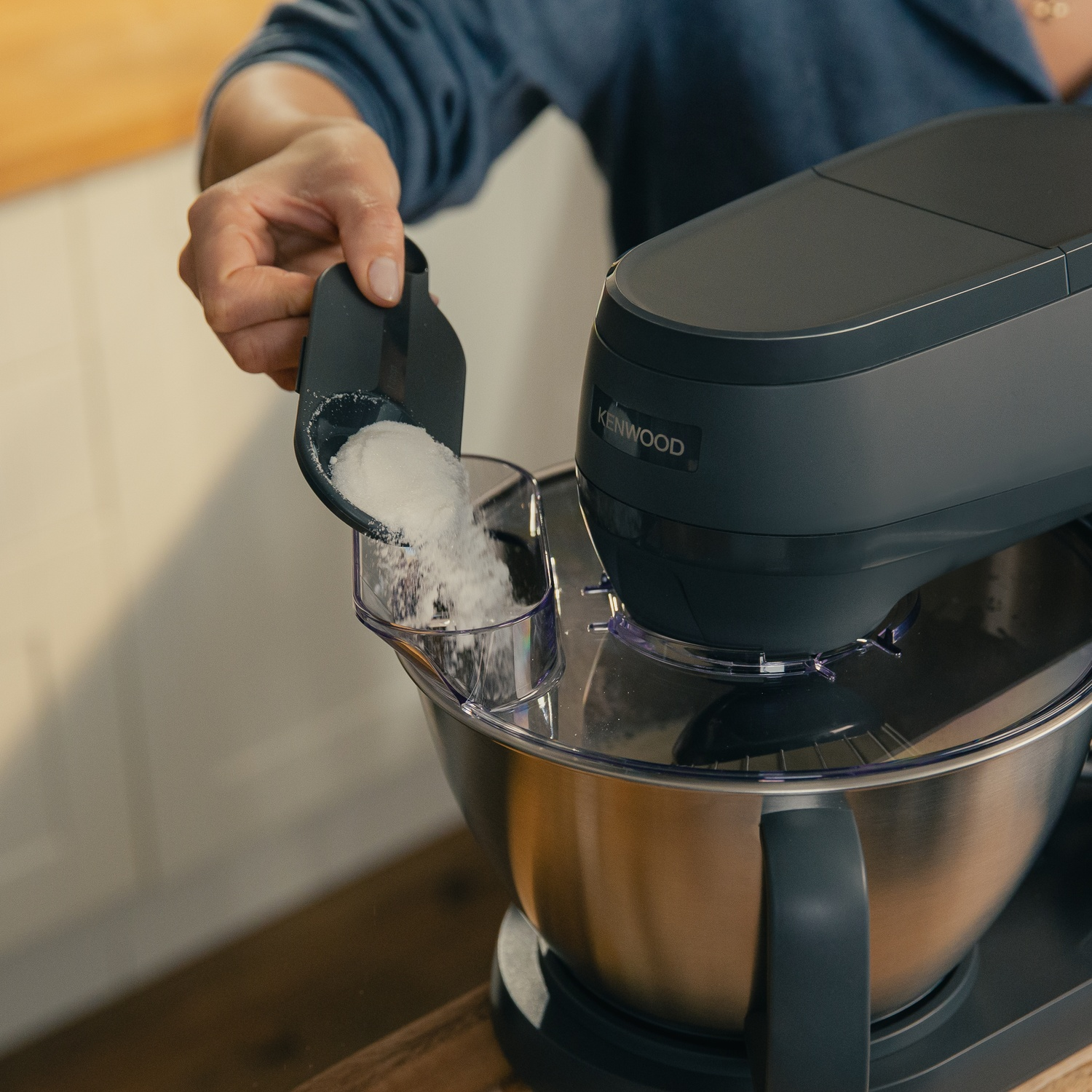 A hand pours white powder into a stand mixer using a scoop.