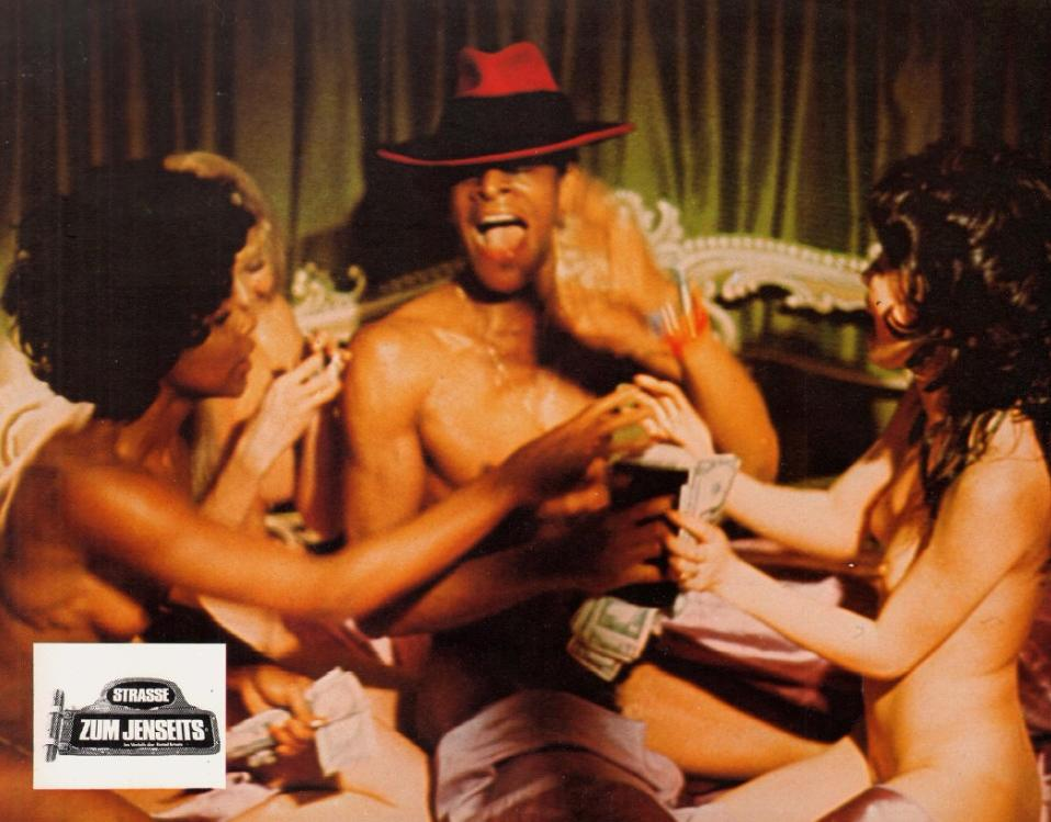 Mann mit rotem Hut bei Frauen, eine gibt Geld. Ein Filmplakat ist sichtbar.