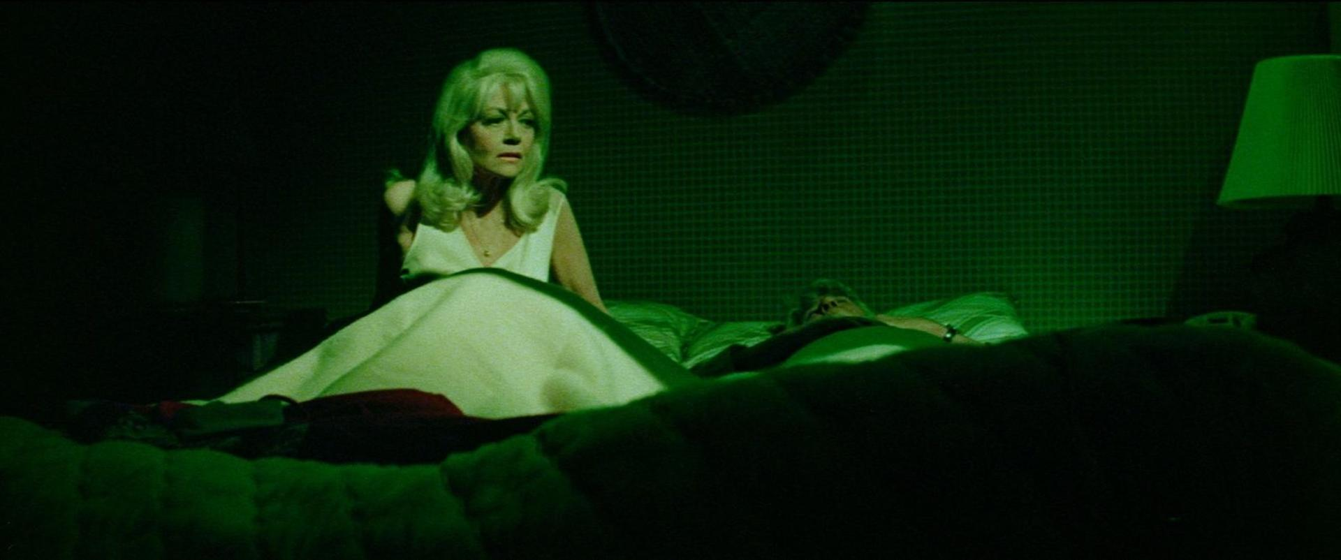 Eine blonde Frau in einem weißen Kleid sitzt im Bett neben einer liegenden Person.