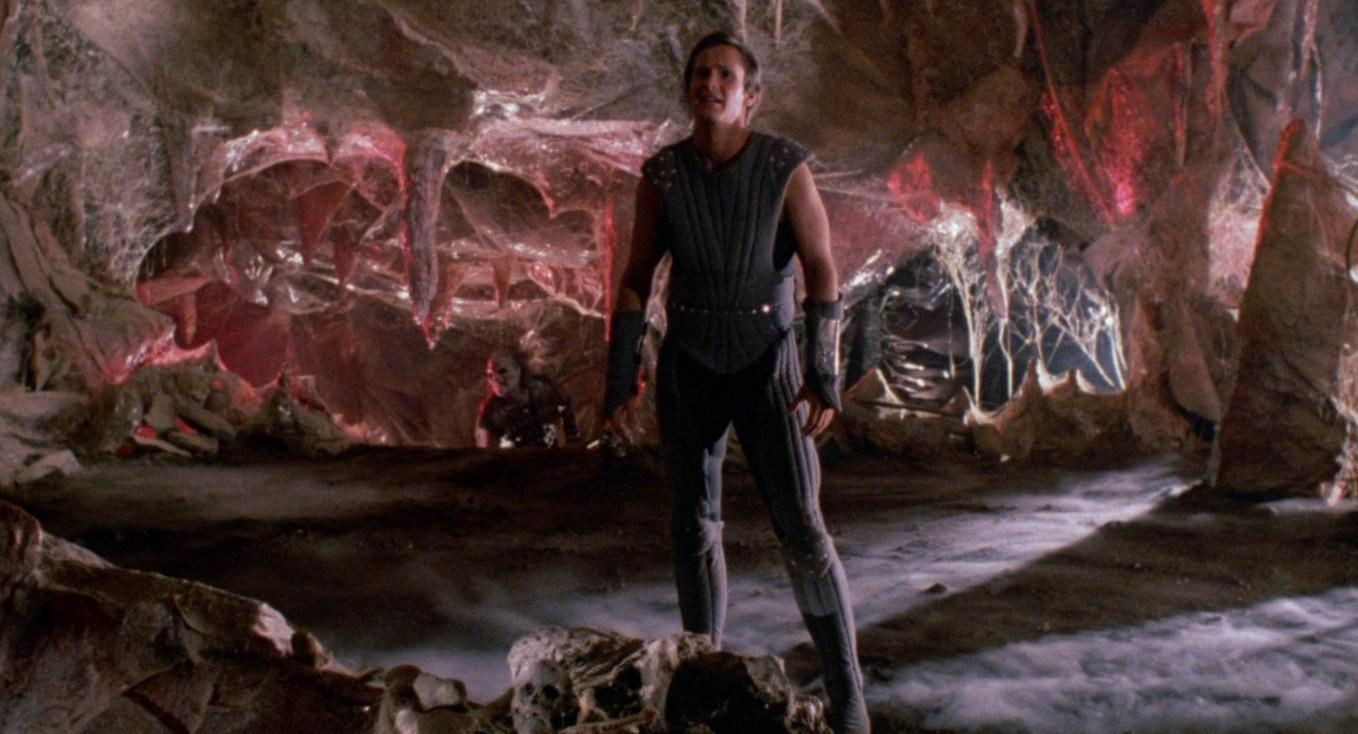 Ein Mann in Rüstung steht in einer Höhle mit spinnennetzartigen Strukturen und rotem Licht.
