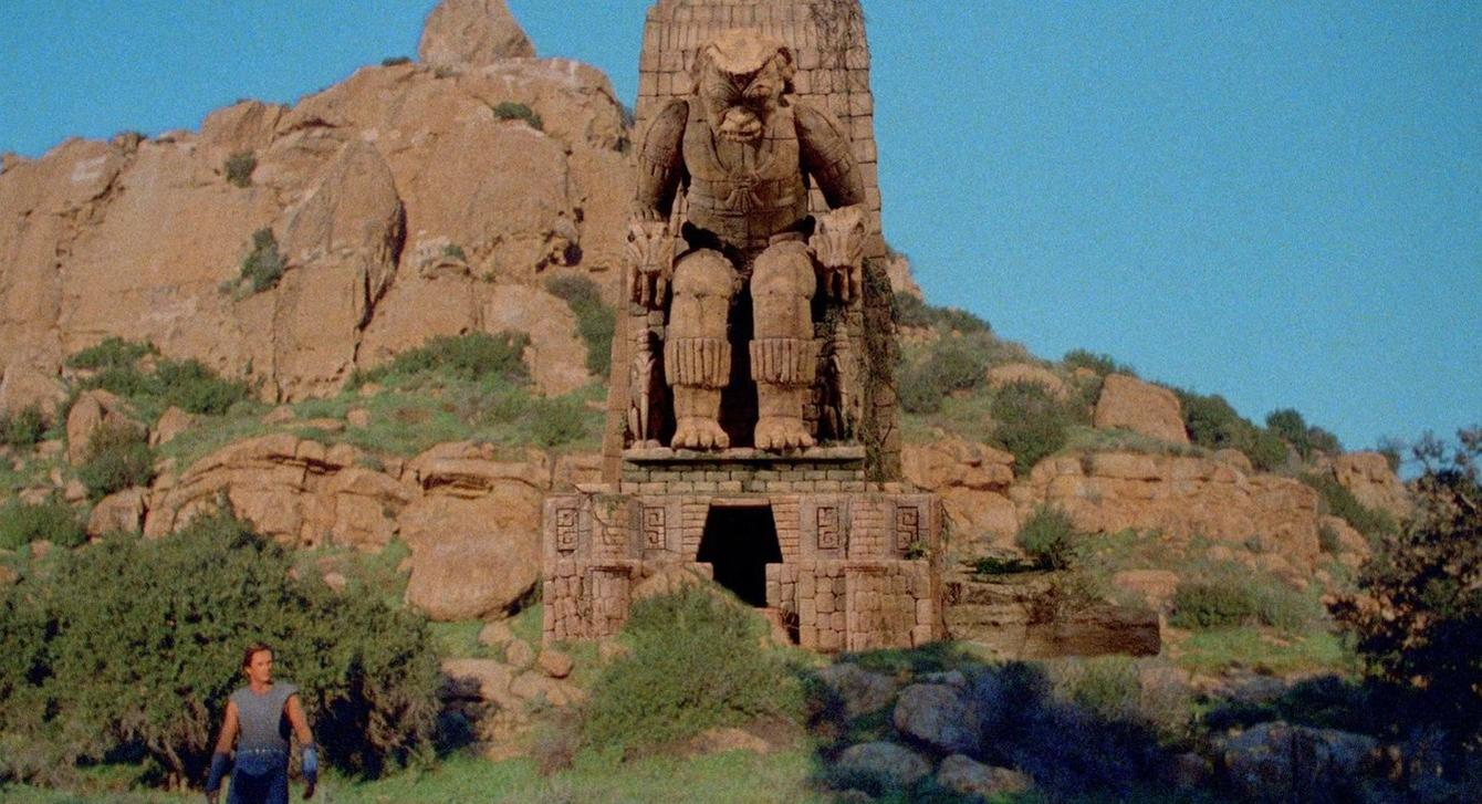 Ein Mann steht vor einer riesigen Steinstatue eines Wesens in einer felsigen Landschaft.