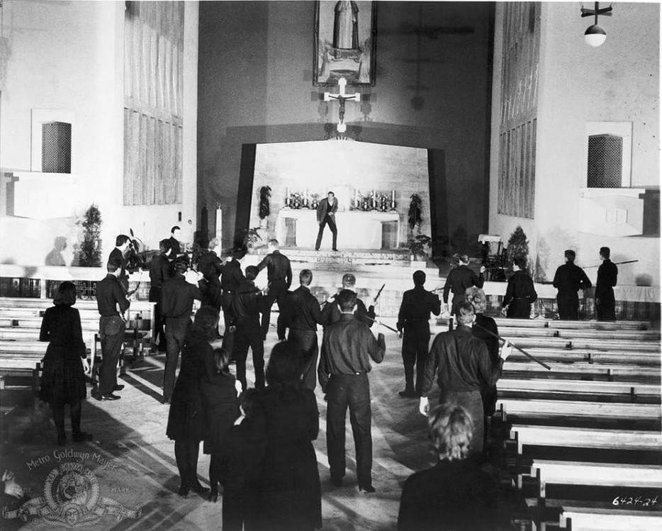 Männer mit Waffen in einer Kirche, eine Person steht auf einer Bühne.