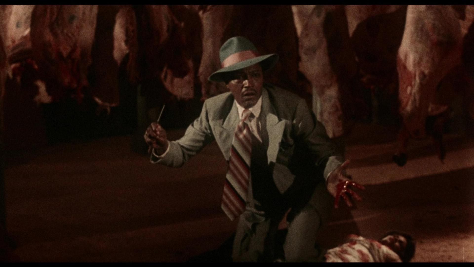 Ein Mann in einem Anzug und Hut kniet und hält einen Dolch. Hängende Tierkadaver im Hintergrund.