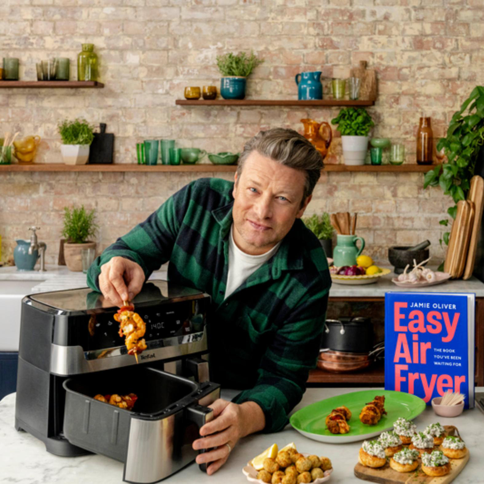 Jamie Oliver präsentiert ein Kochbuch und ein Heißluftfritteusen-Gerät, mit Spießen.