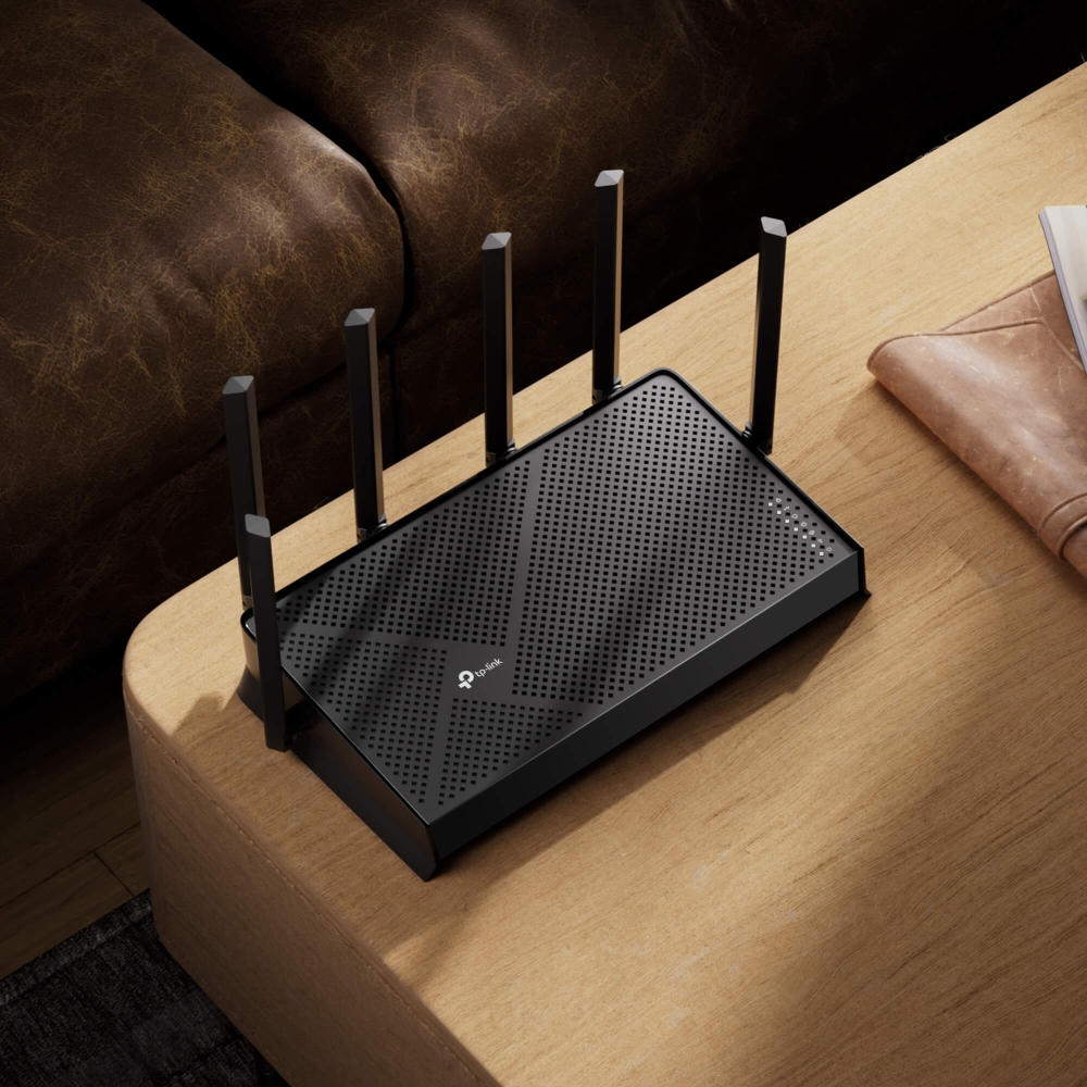 Un router negro con cuatro antenas sobre una mesa de madera junto a un sofá marrón.