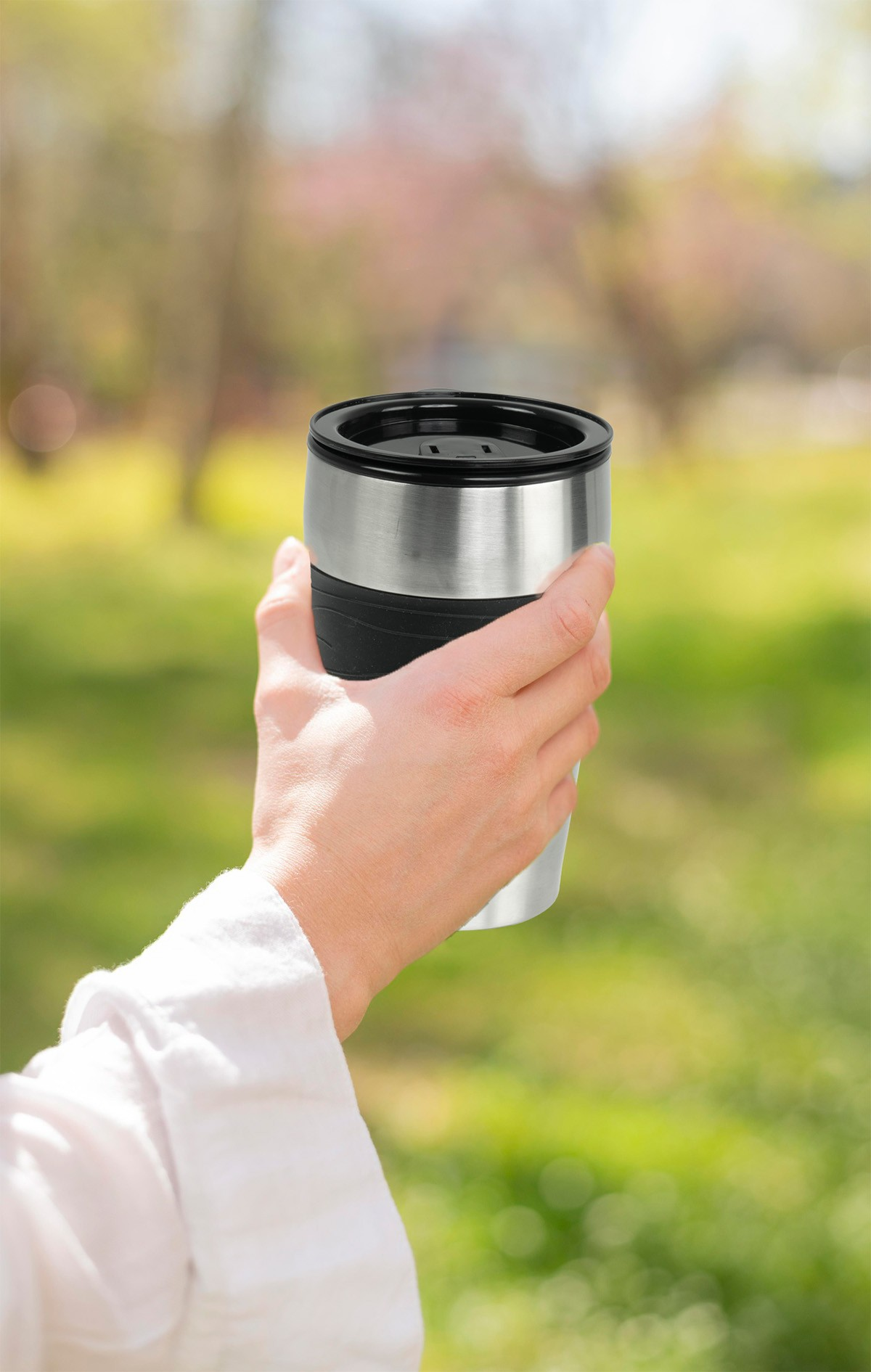 Una mano sostiene una taza de viaje plateada con tapa negra al aire libre.