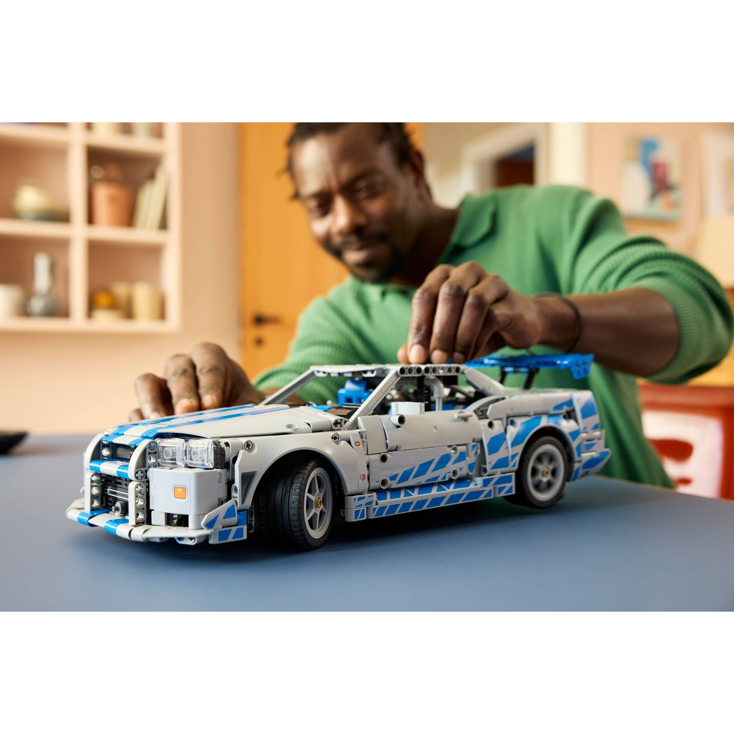 Un uomo costruisce un'auto Lego bianca e blu. È seduto a un tavolo blu.