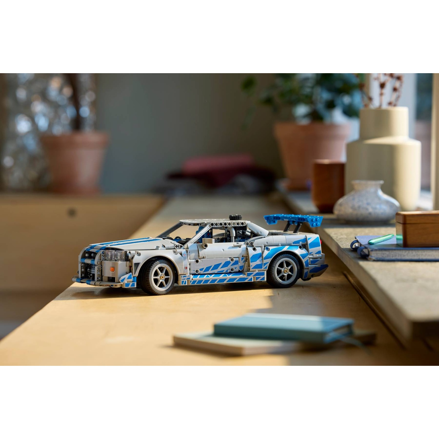 Modellino auto Lego bianco e blu con spoiler su superficie di legno, con piante e vasi sullo sfondo.
