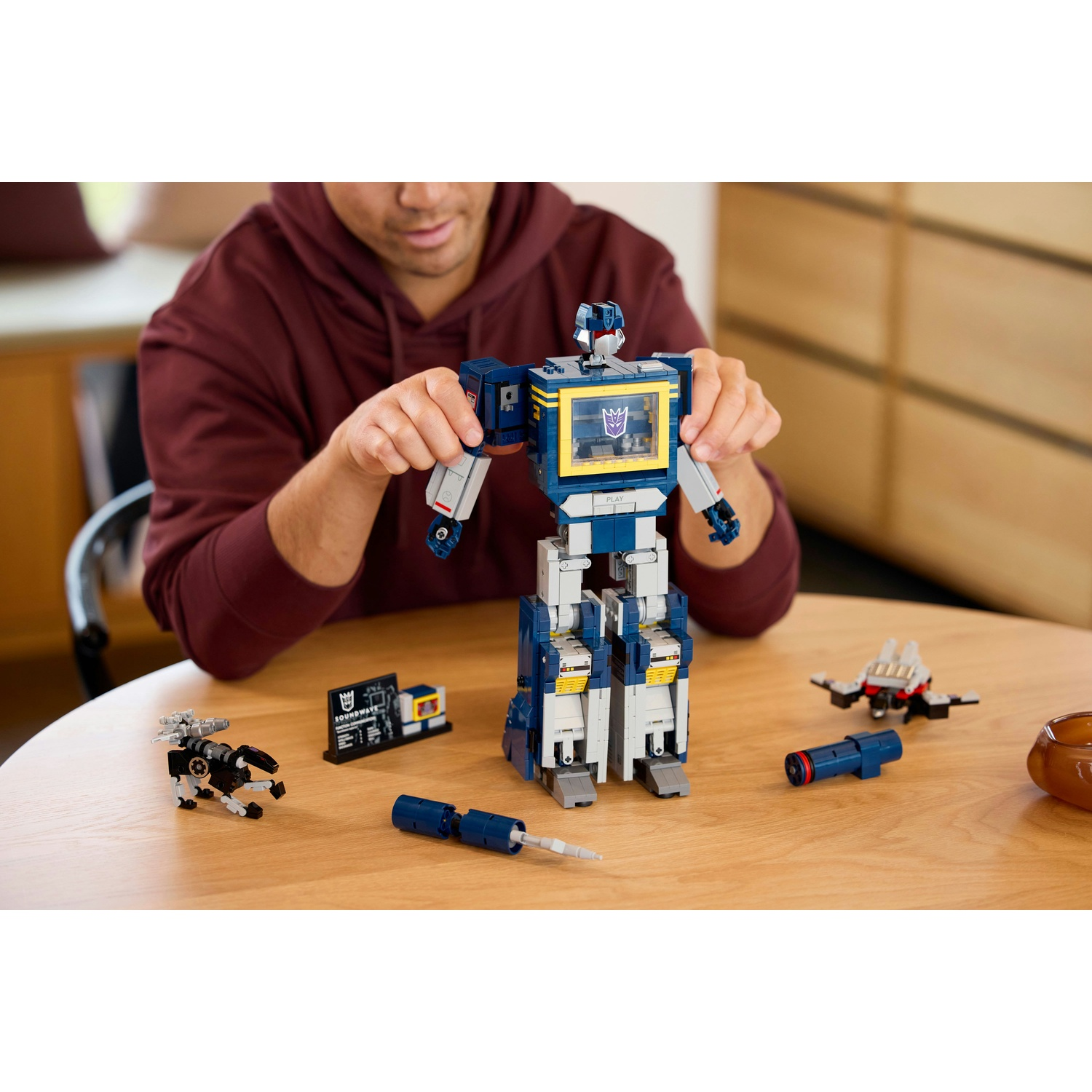 Un uomo assembla un robot Lego Soundwave blu e bianco su un tavolo di legno con accessori.