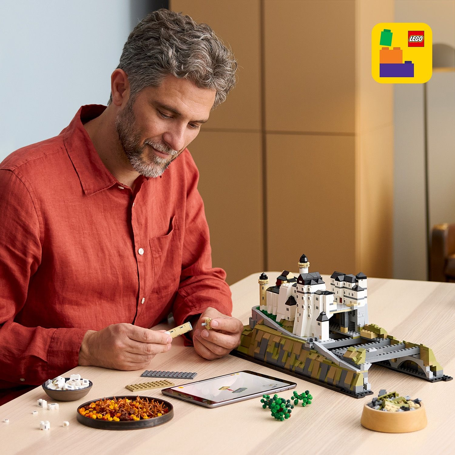 Uomo in camicia rossa costruisce castello Lego su tavolo, con pezzi e tablet.