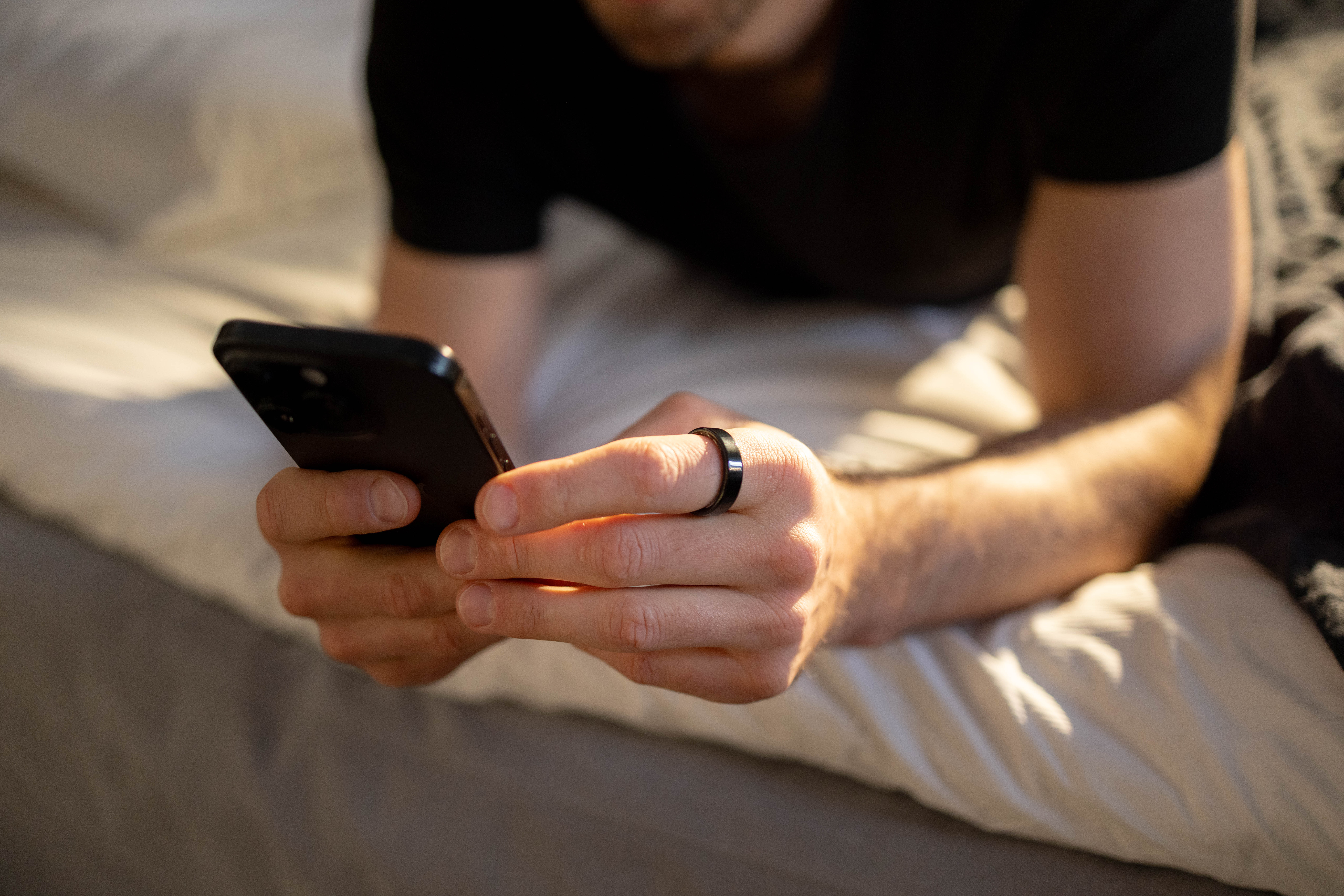 Eine Person in einem schwarzen T-Shirt liegt auf einem Bett und hält ein Smartphone.